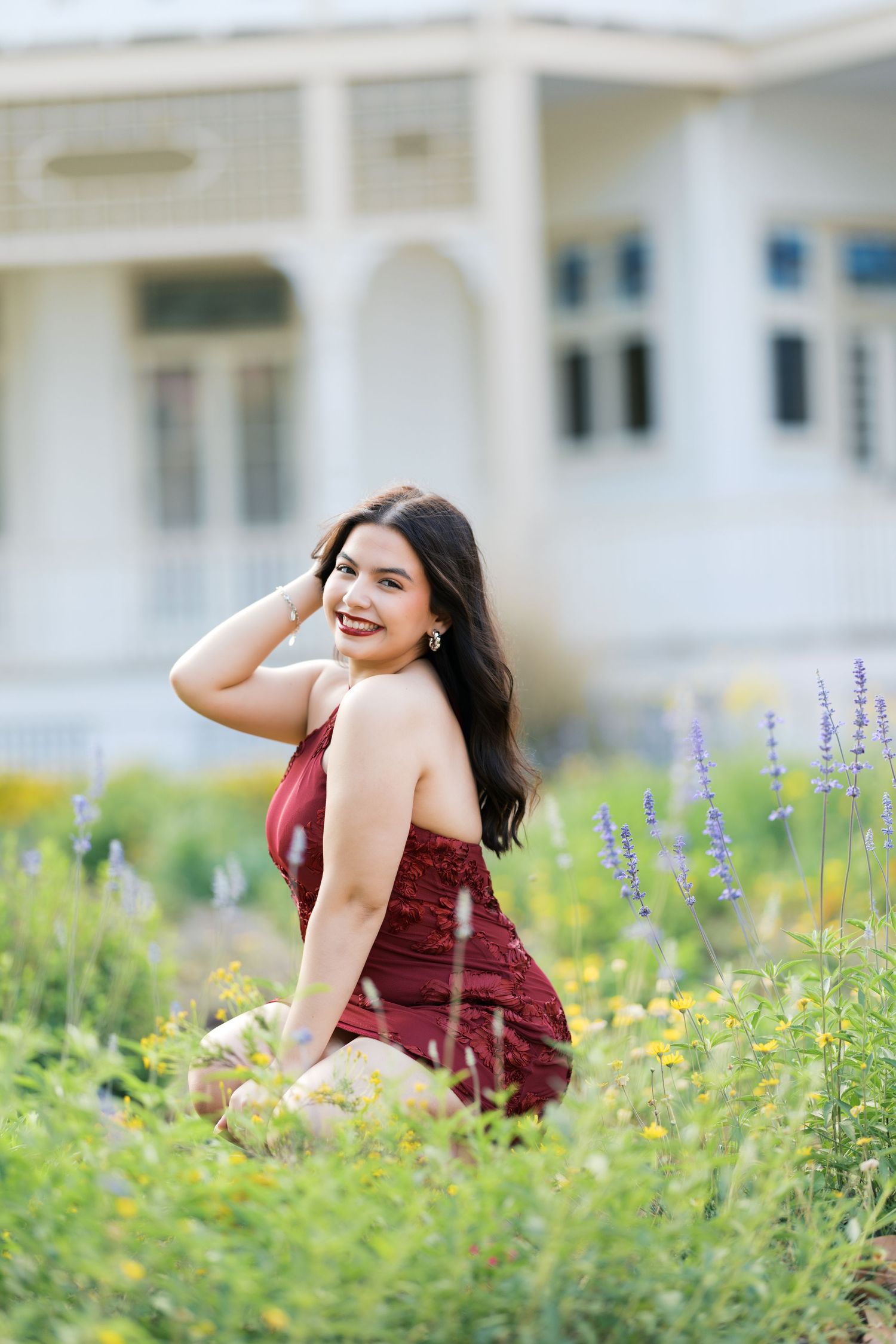 Young woman in red dress sitting among wildflowers in a garden, smiling over her shoulder.