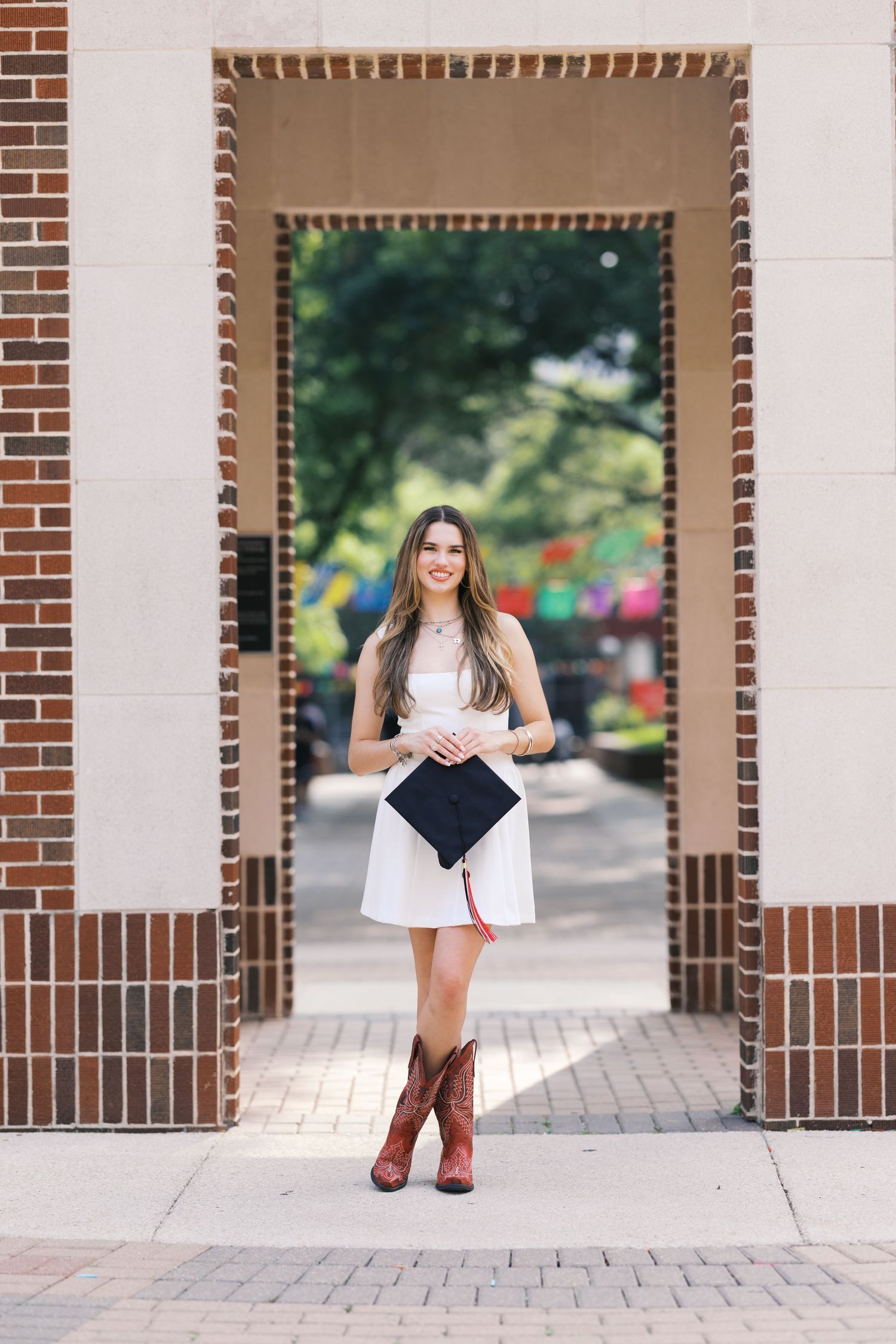 Young woman in white dress and cowboy boots holds graduation cap while standing in a brick archway on campus.