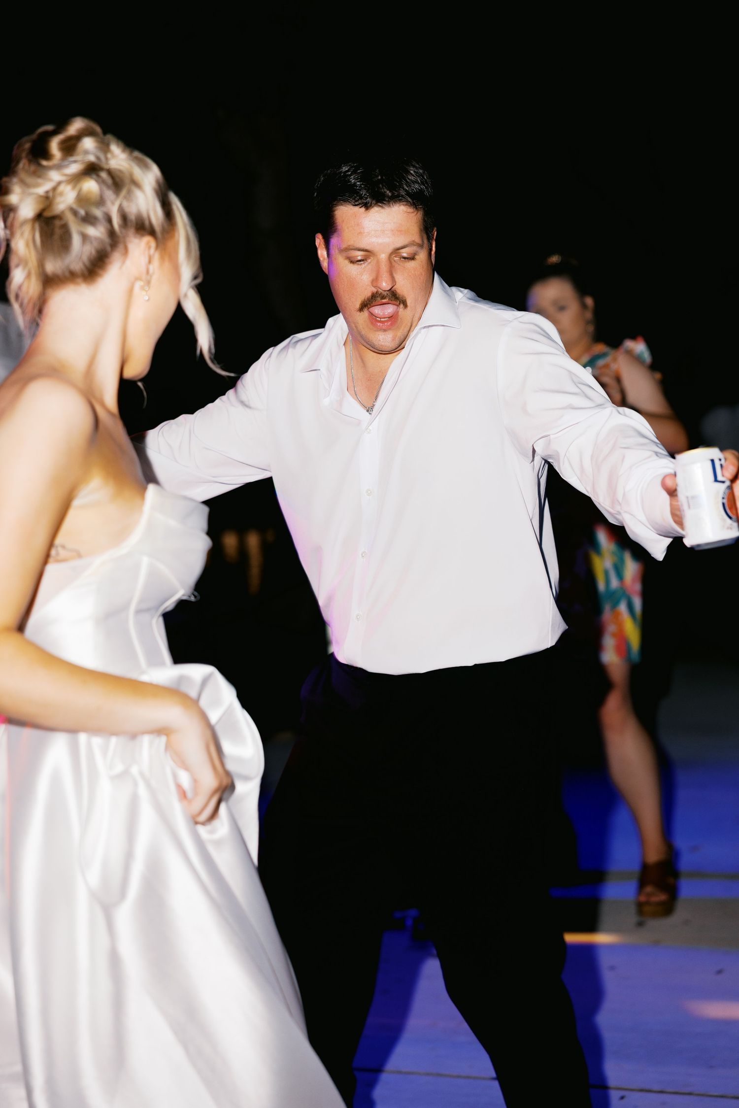 A man with a mustache in a white shirt dances energetically with a bride in a white gown at a wedding reception.
