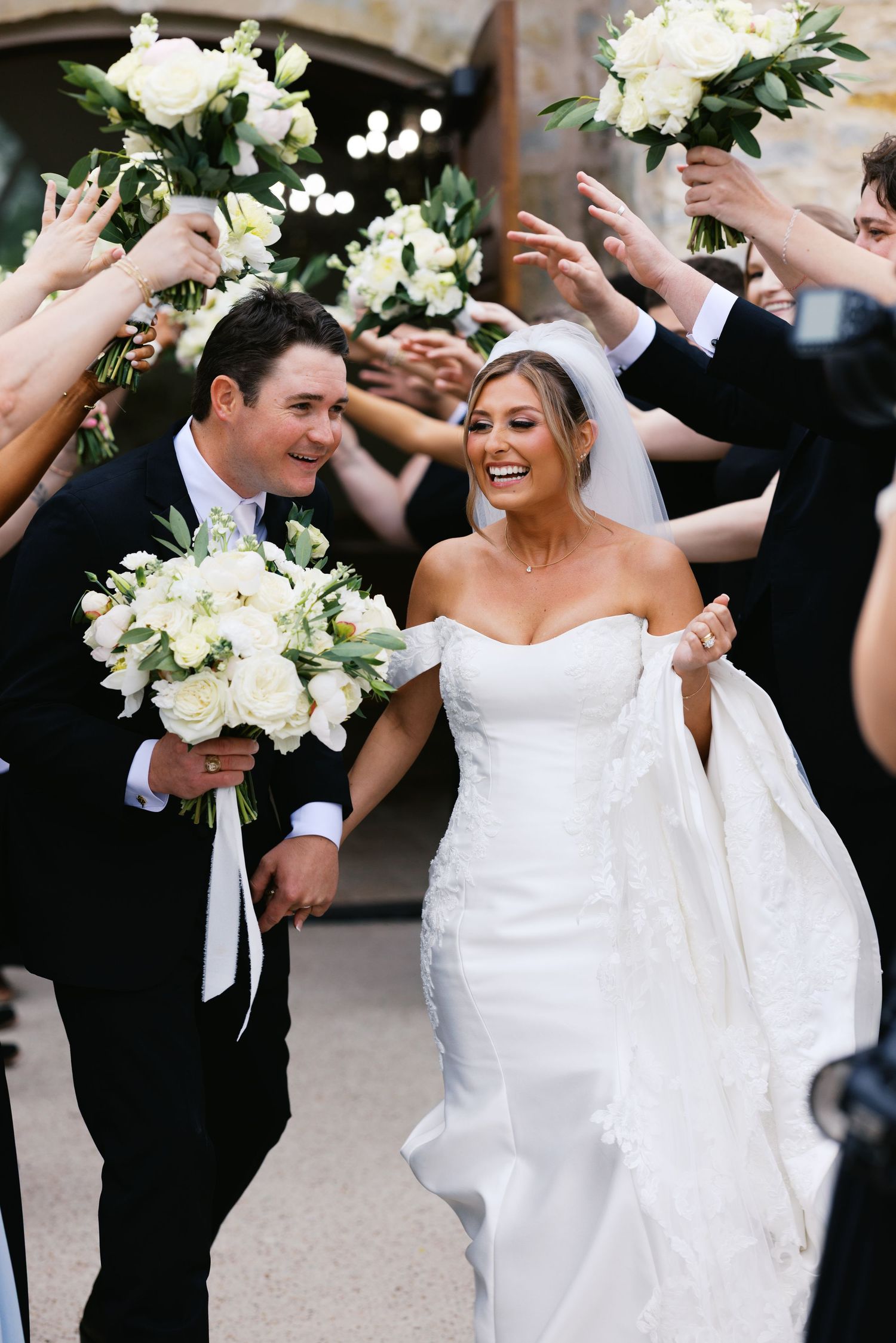 Laughing bride and groom exit church as wedding party raises bouquets in celebratory arch overhead.