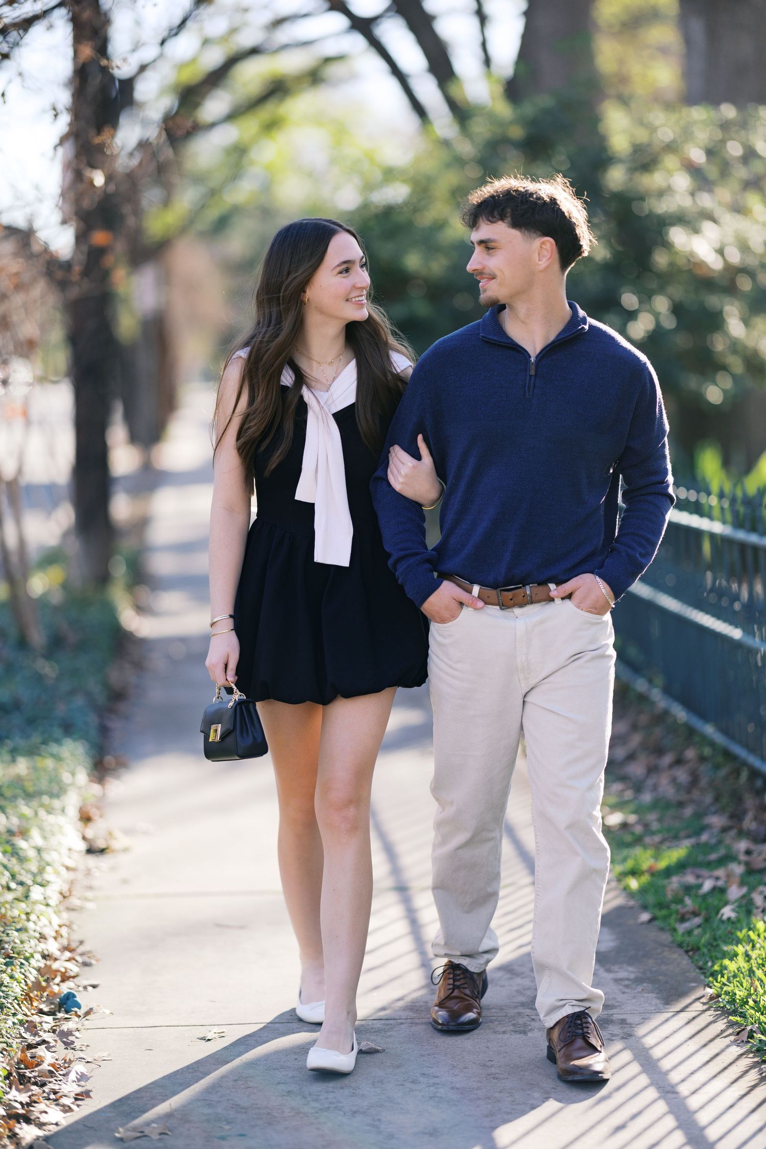 A young couple walks arm in arm on a sunny sidewalk, smiling at each other, dressed in smart casual attire.