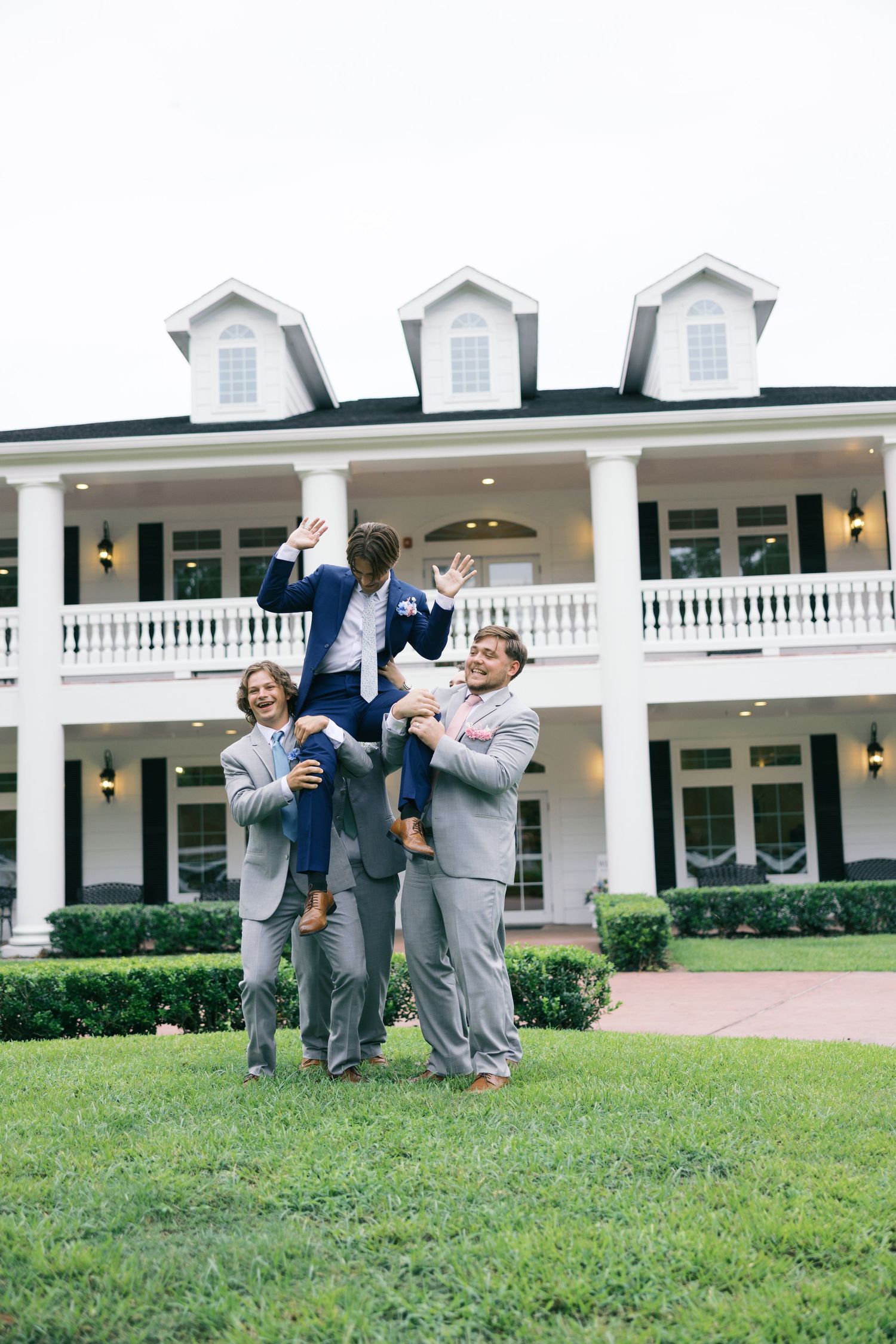 Groomsmen in gray suits celebrating on lawn in front of white colonial mansion.