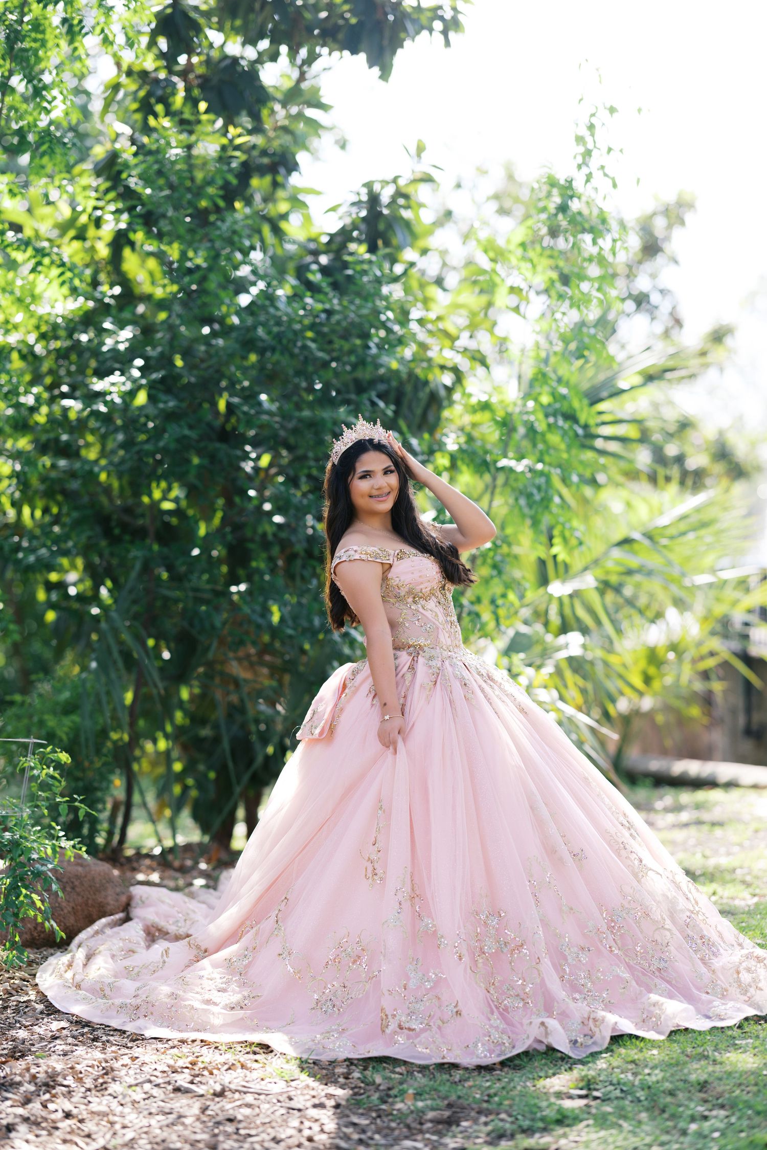 Young woman in elegant pink gold quinceañera gown with tiara poses gracefully outdoors among green trees.