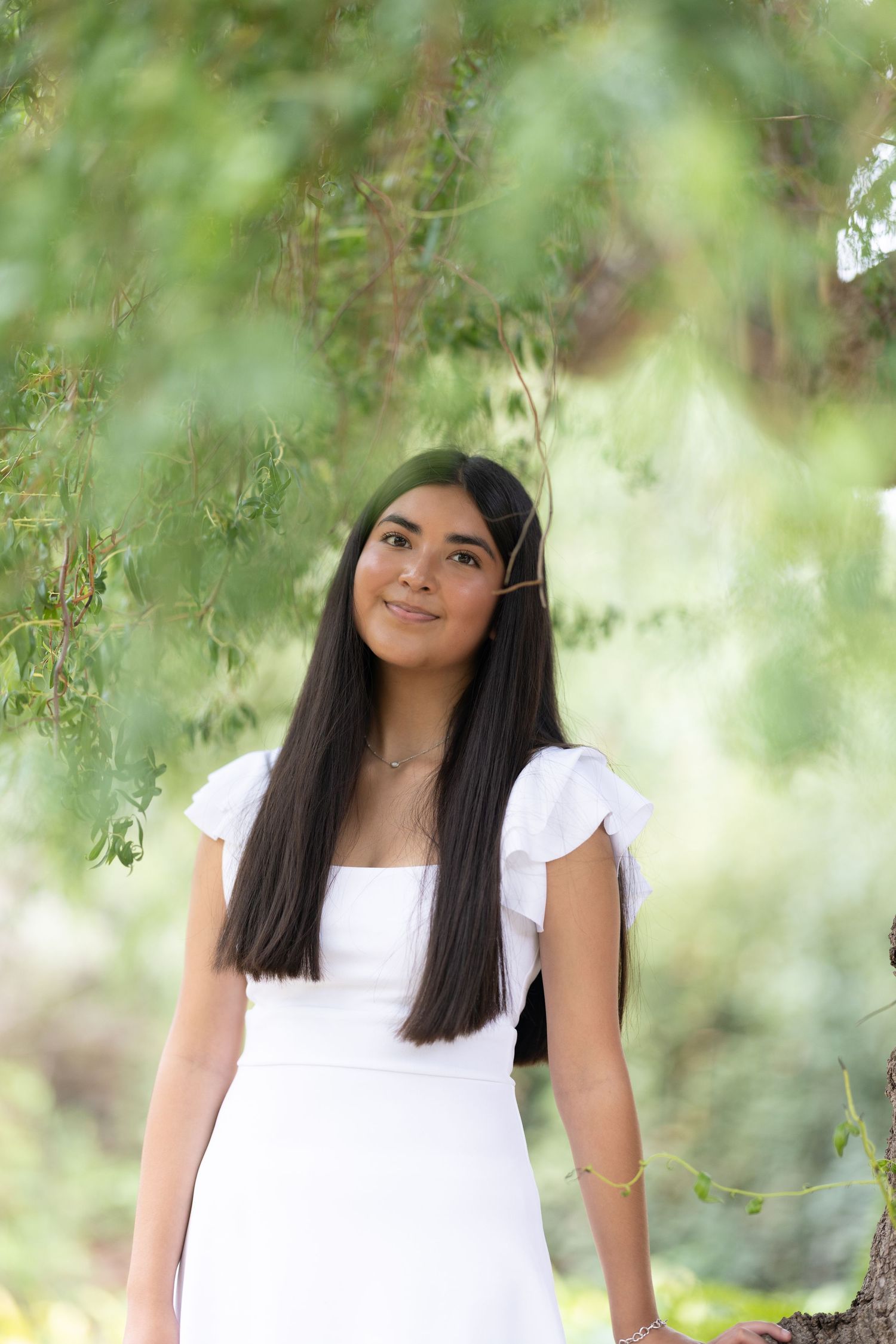 Teen girl in white dress with flutter sleeves smiling under weeping willow tree branches outdoors.