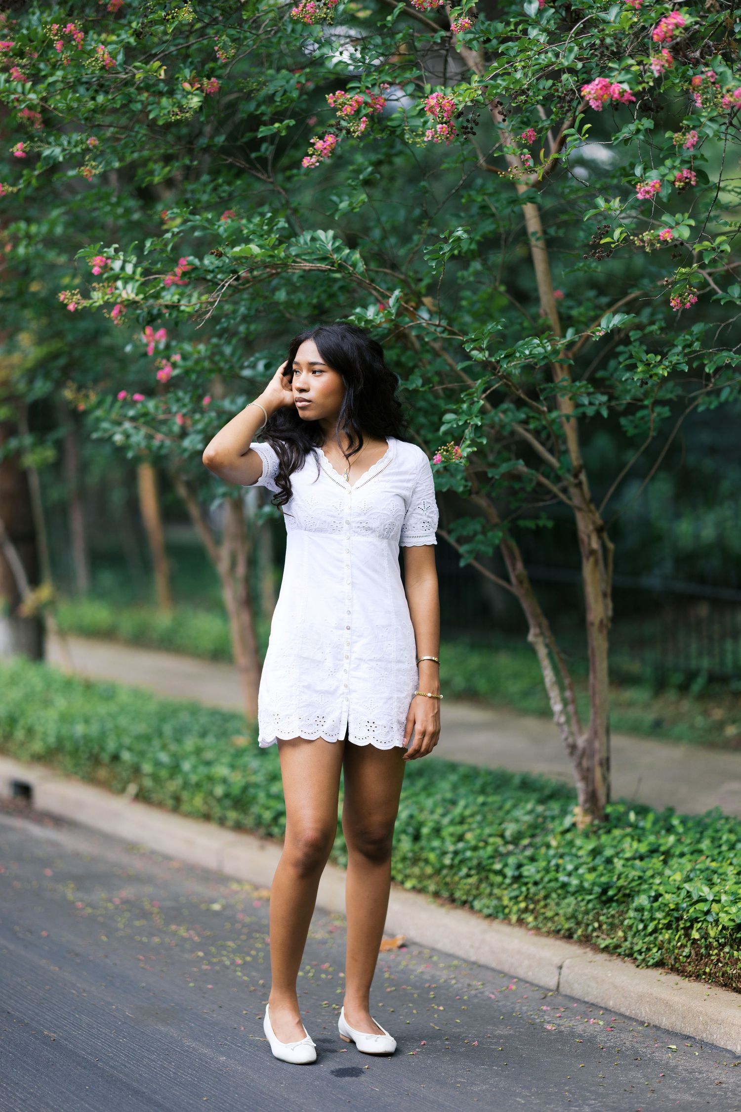 Young woman in white eyelet dress stands on tree-lined path with blooming pink crepe myrtle trees behind her.