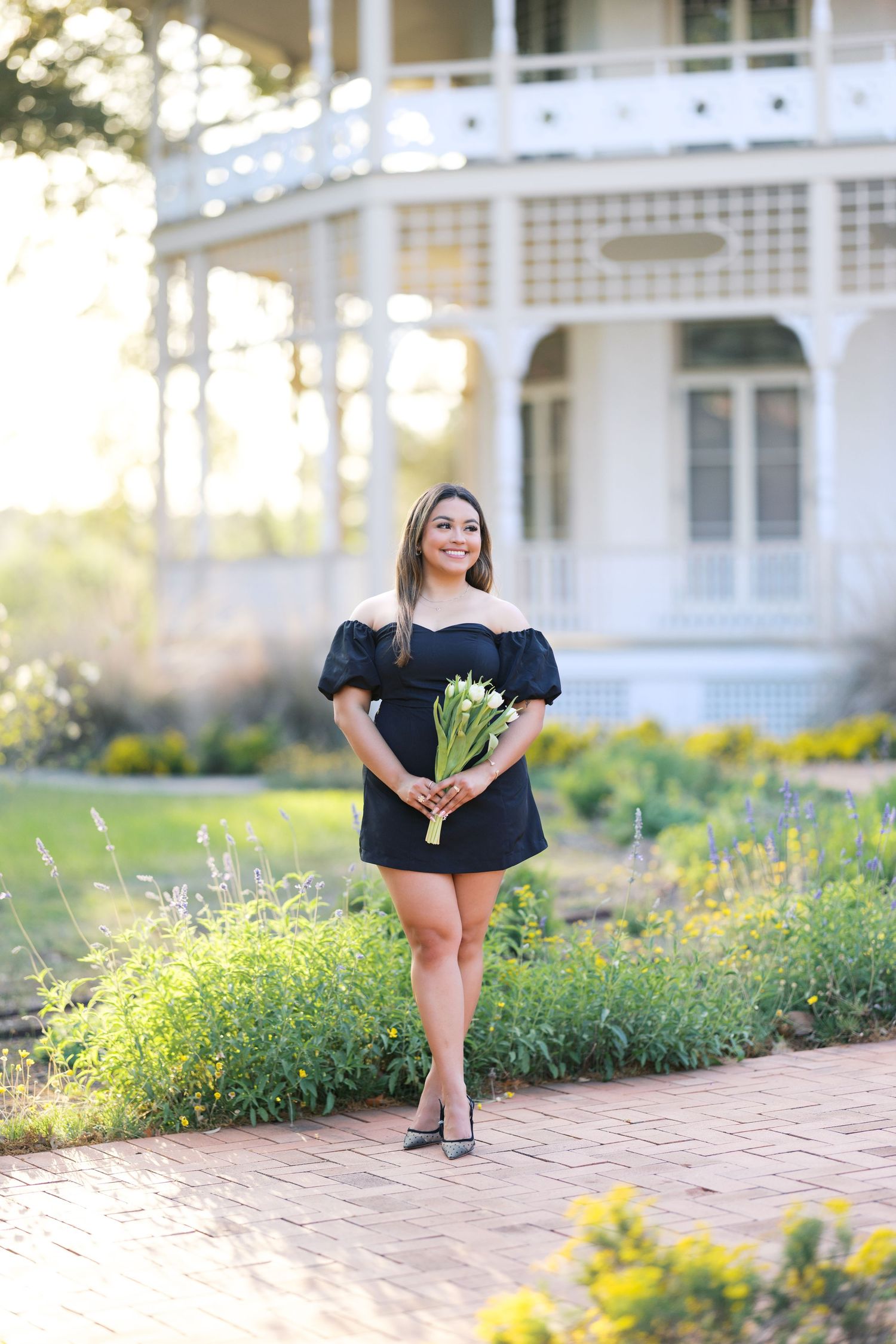 Young woman in black off-shoulder mini dress holds white tulips, posing on brick path near white gazebo garden.