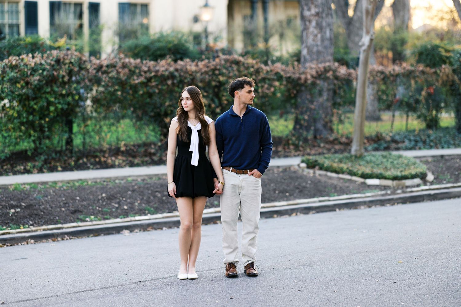 A young couple holds hands while walking on a tree-lined street, dressed in casual elegant attire.