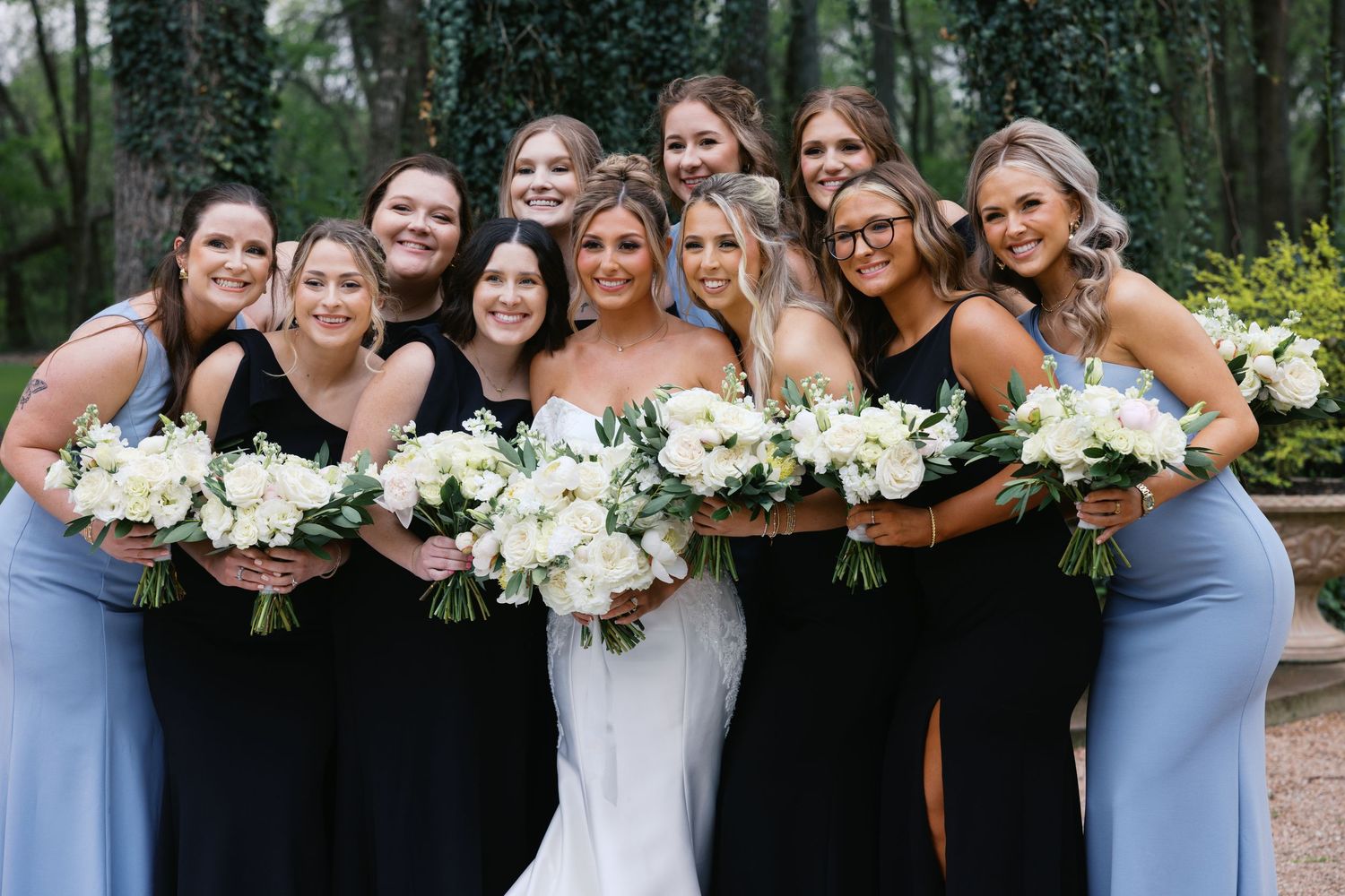 Bride in white gown poses with bridesmaids in black and blue dresses holding white floral bouquets outdoors.