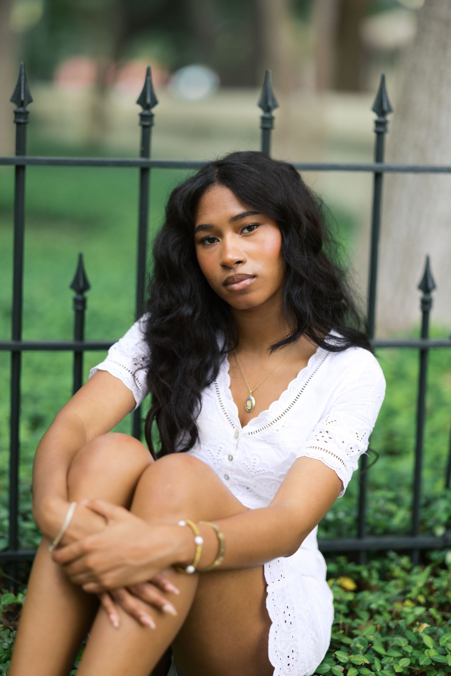 Young woman in white mini dress sitting gracefully beside black iron fence on green grass.