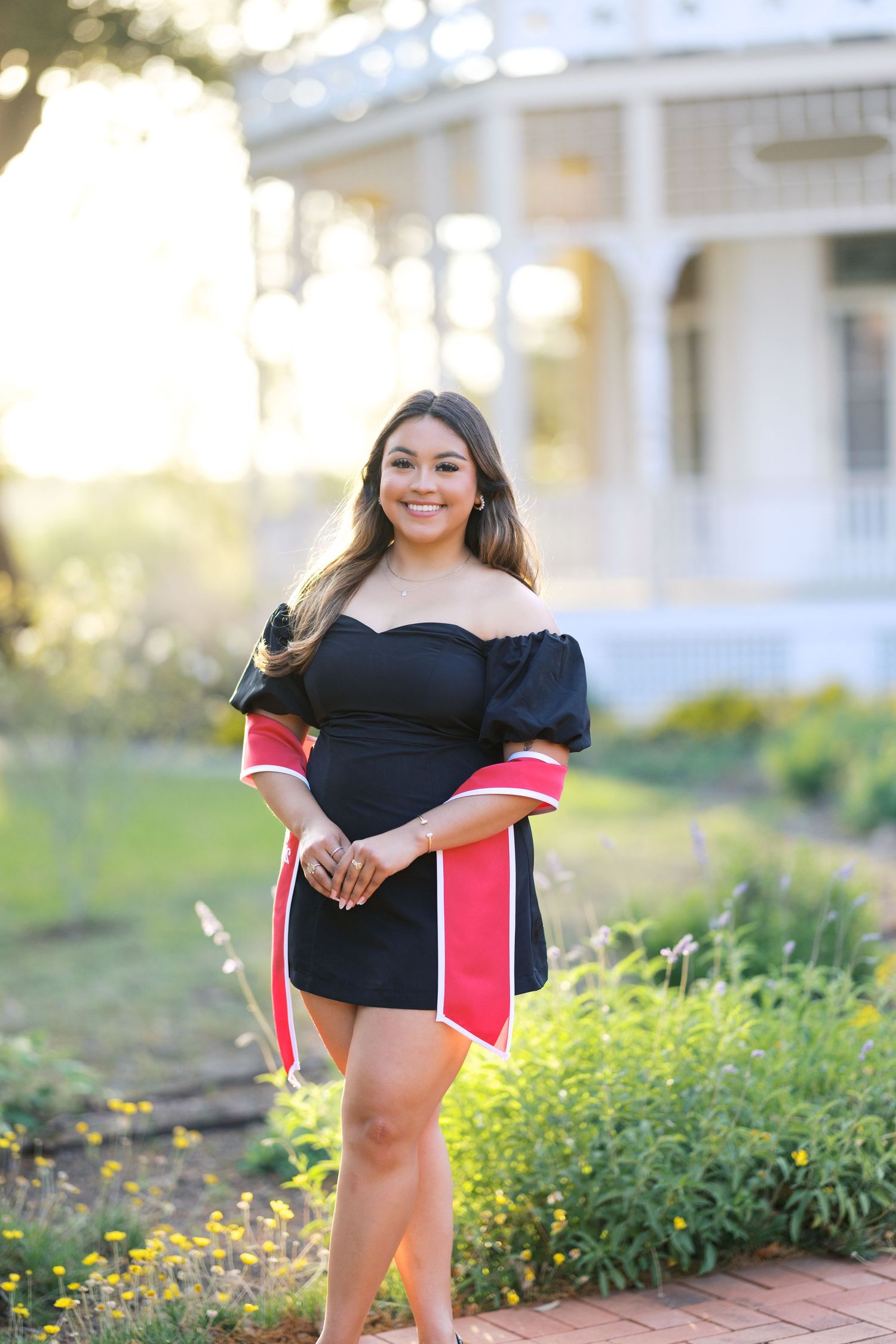 Young woman in black off-shoulder dress with red accents poses outdoors near a white Victorian house.