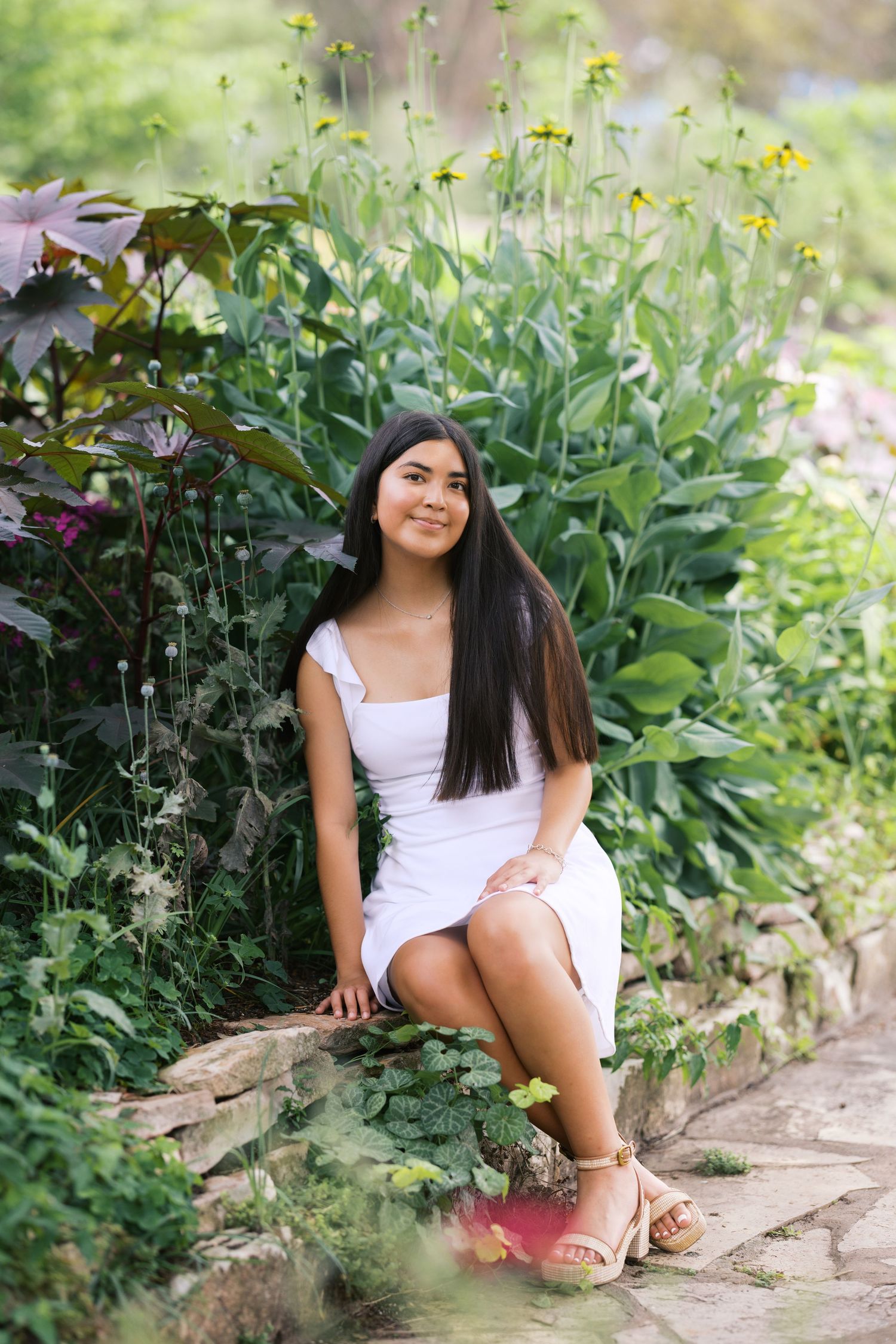 Young woman in white dress sitting on stone wall surrounded by lush green garden with yellow flowers.