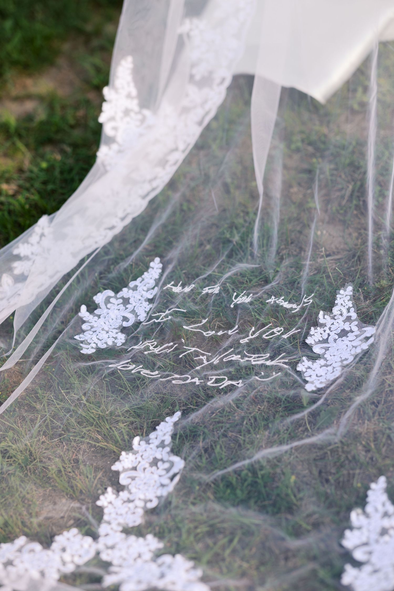 Close-up of a lace-trimmed wedding veil with handwritten script text laid on green grass.
