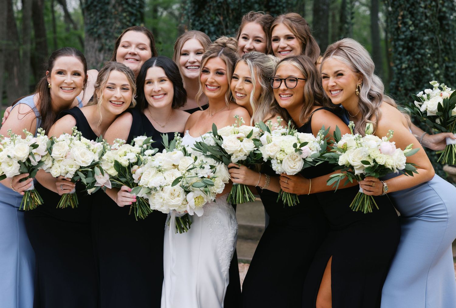 Bride surrounded by bridesmaids in black and blue dresses holding white floral bouquets, smiling outdoors.