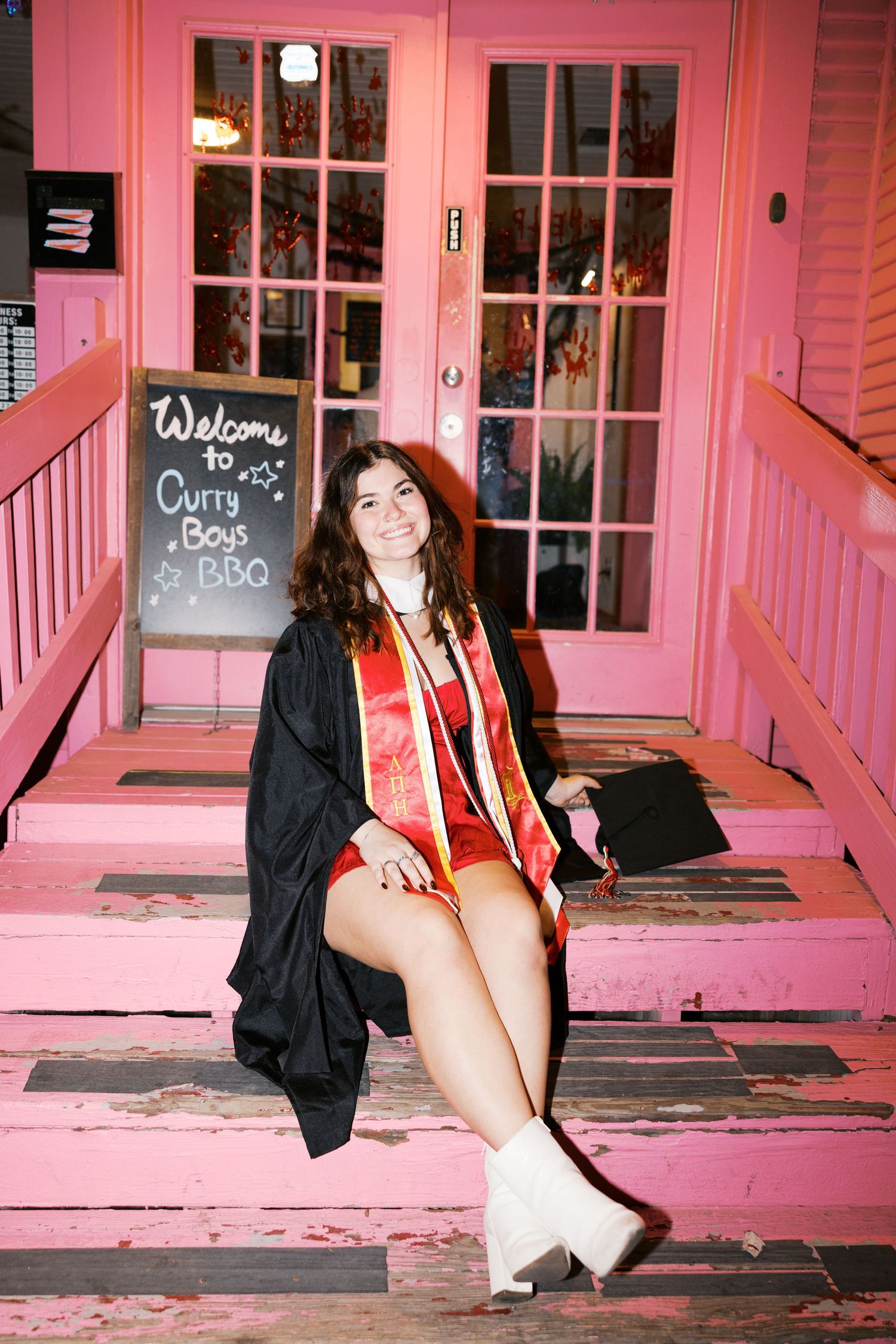 Graduate in academic regalia sitting on pink porch steps near welcome sign.