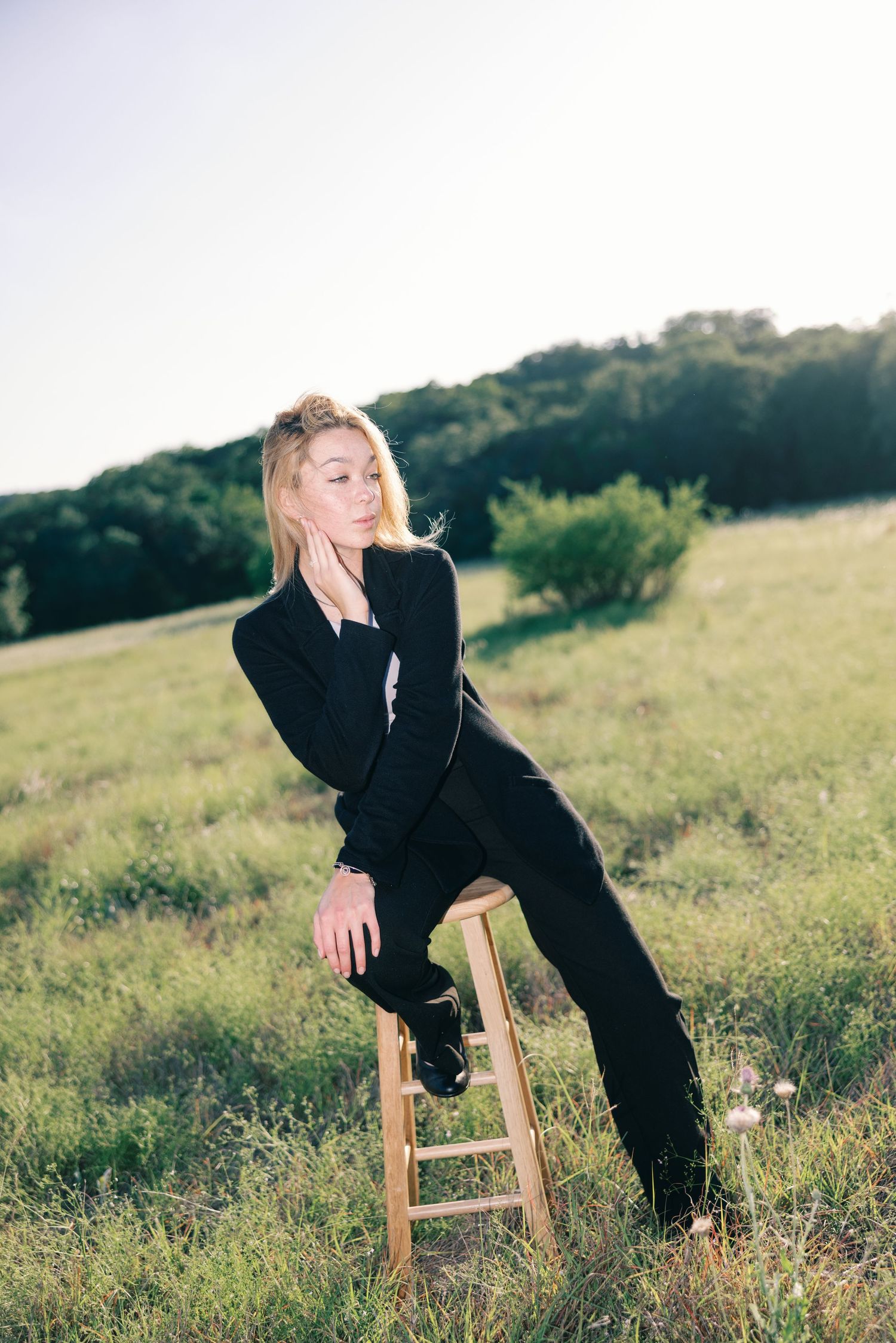 Person in black outfit posing artistically with wooden stool in sunny meadow.
