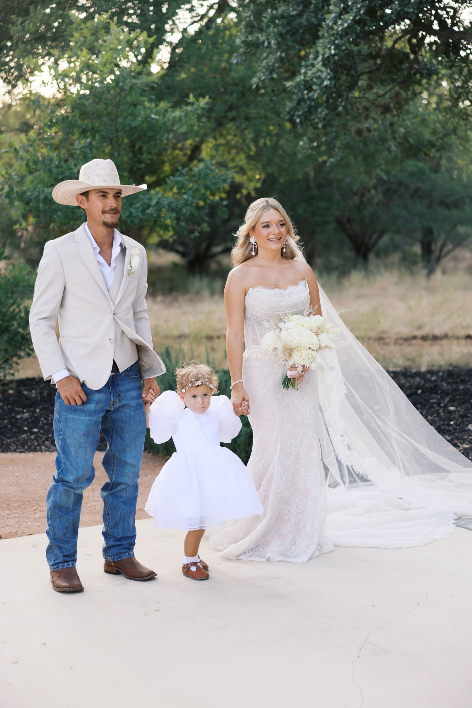 Rustic country wedding photos with a white dress, cowboy attire, and flowing veil against trees.