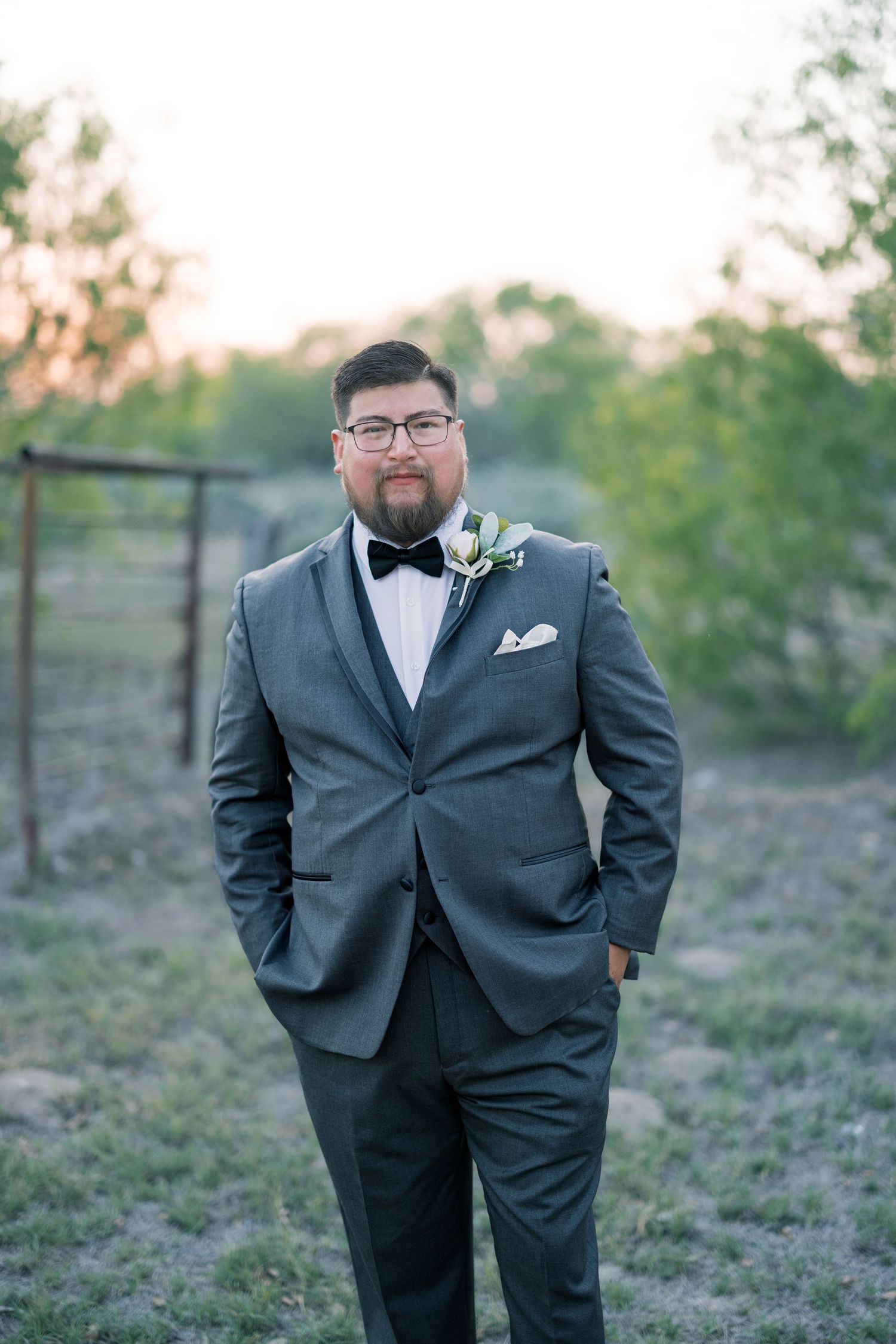A groom in a classic grey suit and bowtie poses outdoors during golden hour.