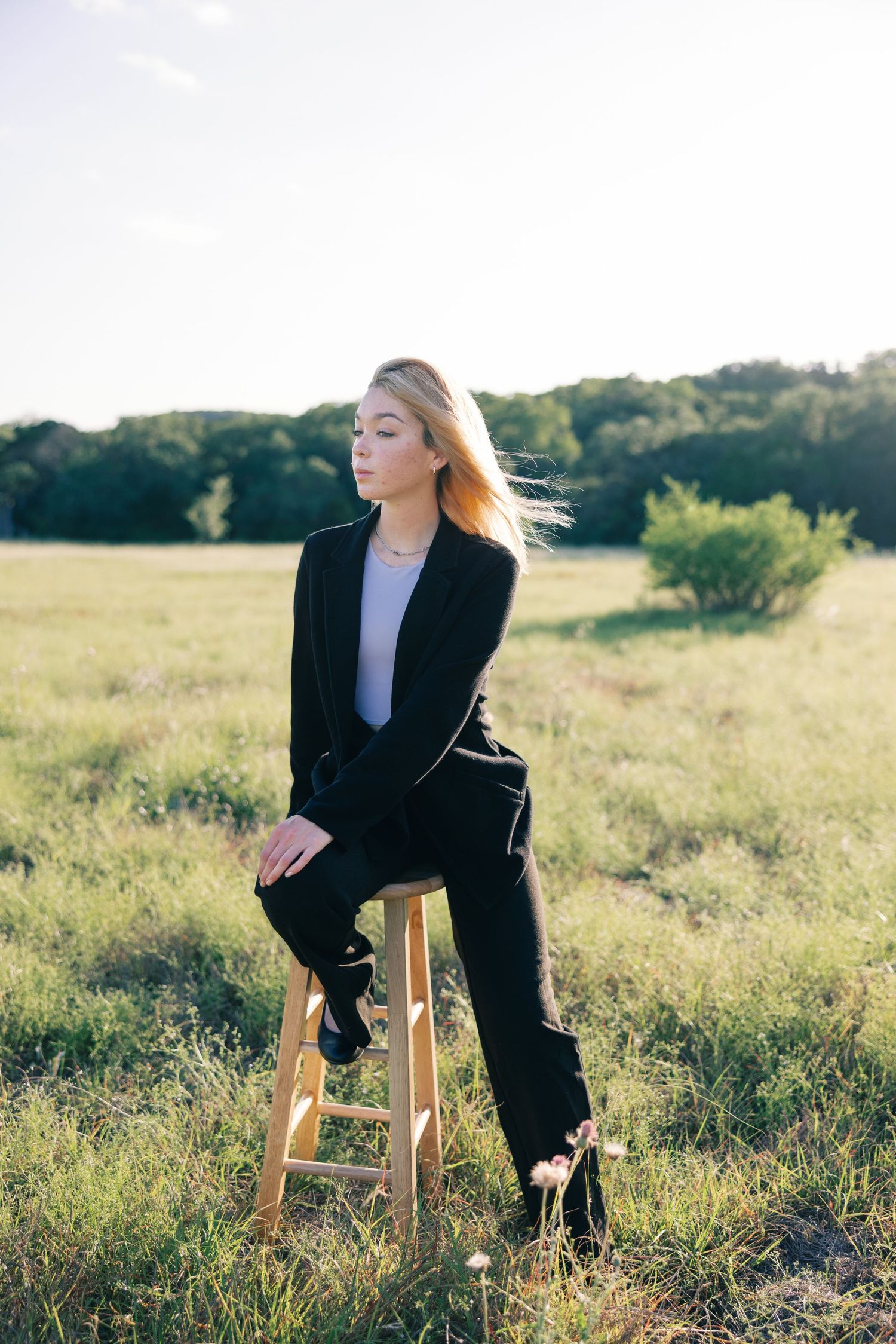 Stylish black suit ensemble photographed on wooden stool in natural meadow setting.