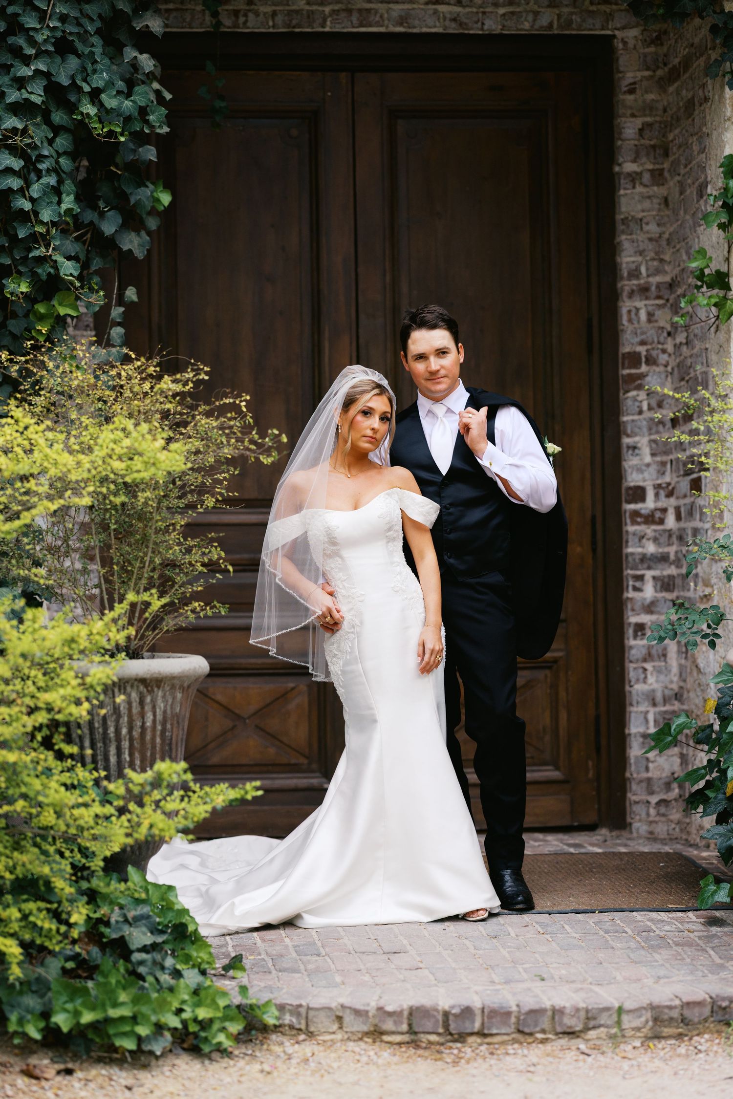 Bride and groom posing together in front of rustic wooden doors surrounded by ivy and lush greenery.