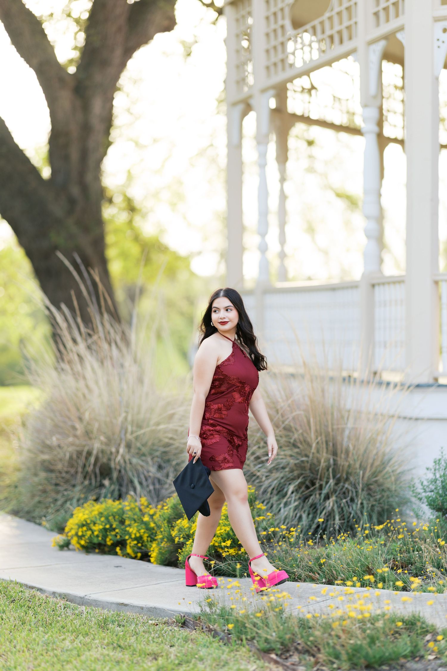 Woman in burgundy dress and pink heels walking on a garden path near a white gazebo with yellow flowers.