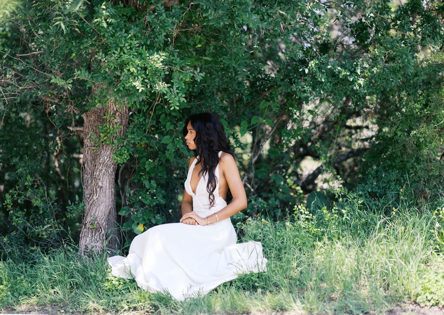 Woman in flowing white dress sitting serenely in tall green grass beneath shady trees.