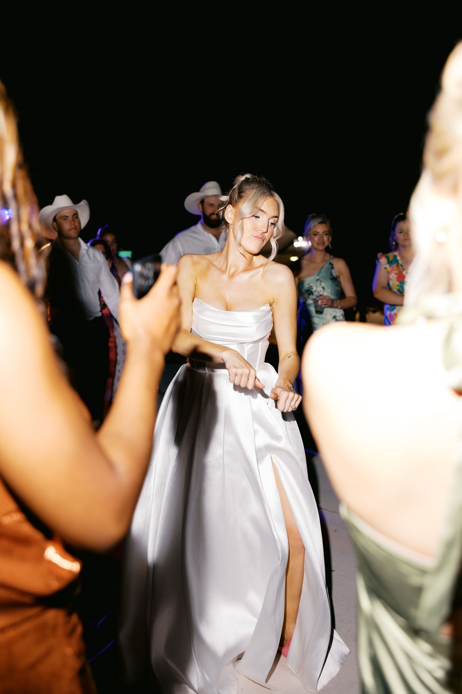 Bride in white strapless ball gown dances energetically at outdoor wedding reception at night.