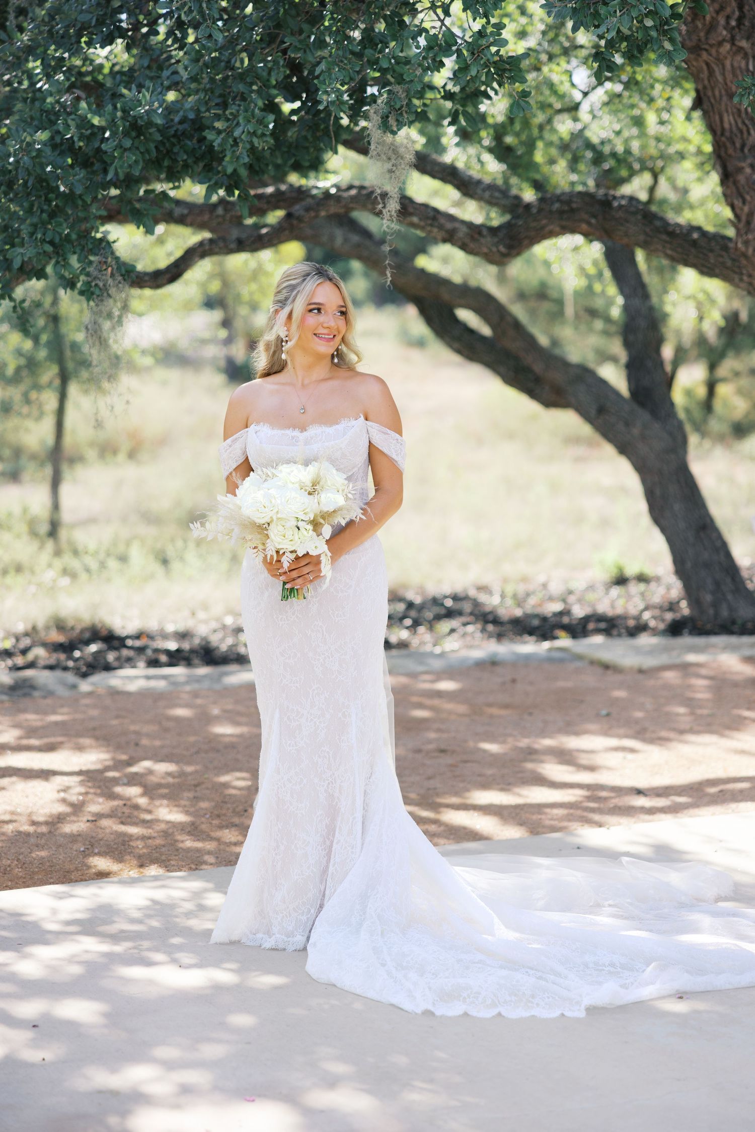 Elegant bride in off-shoulder lace gown with long train poses under a large oak tree.