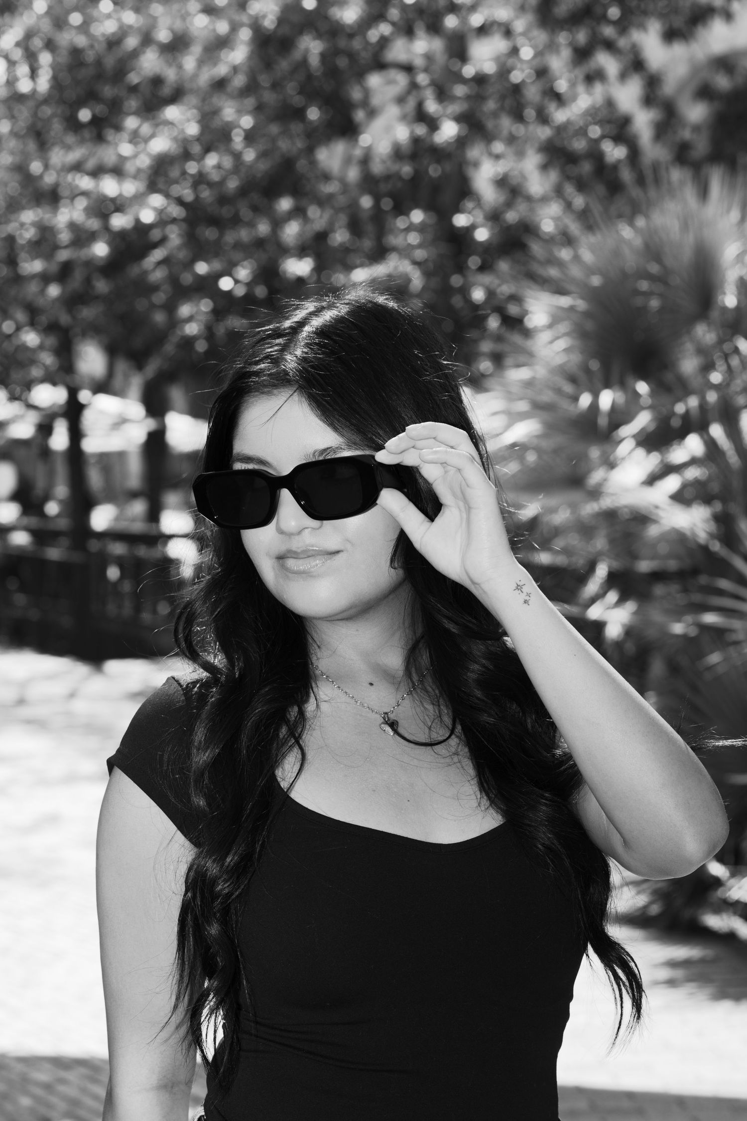 Black and white portrait of person in sunglasses and black top against lush garden background.