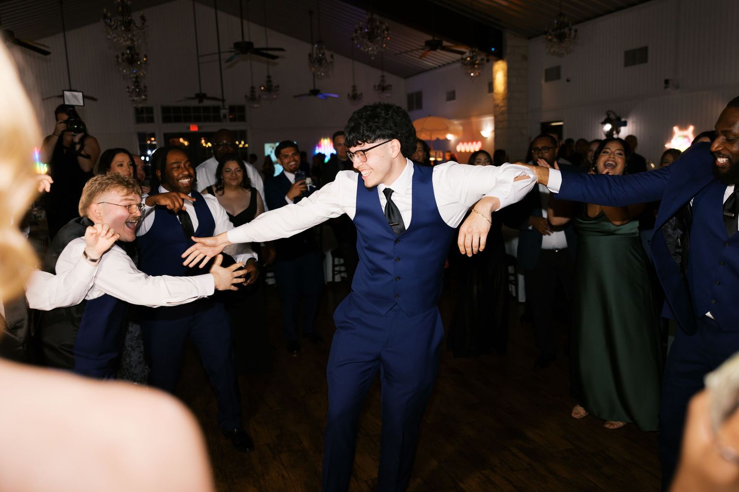 Guests in navy suits dancing energetically at a wedding reception in a warmly lit venue.