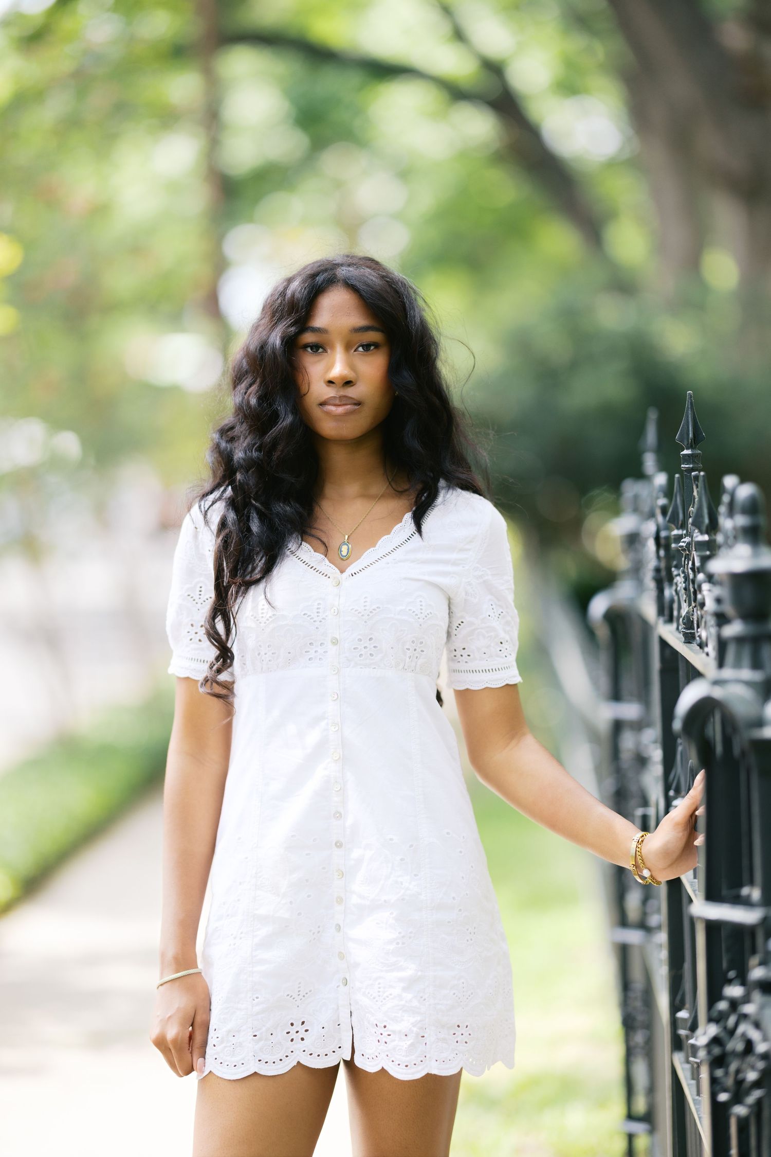 Young Black woman in white eyelet dress standing by iron fence on tree-lined sidewalk.