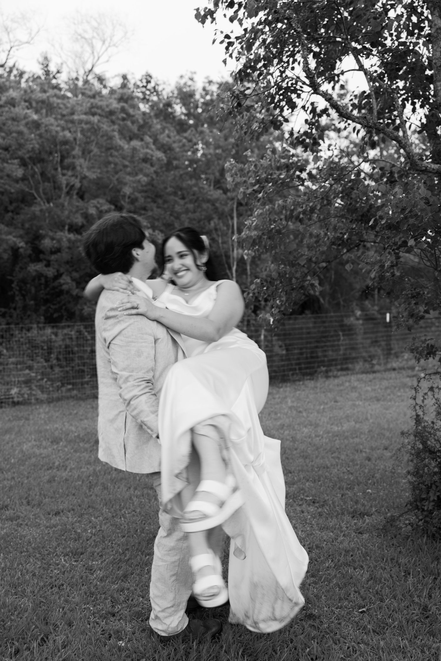Black and white photo of groom lifting bride in wedding dress outdoors on grass with trees in background.