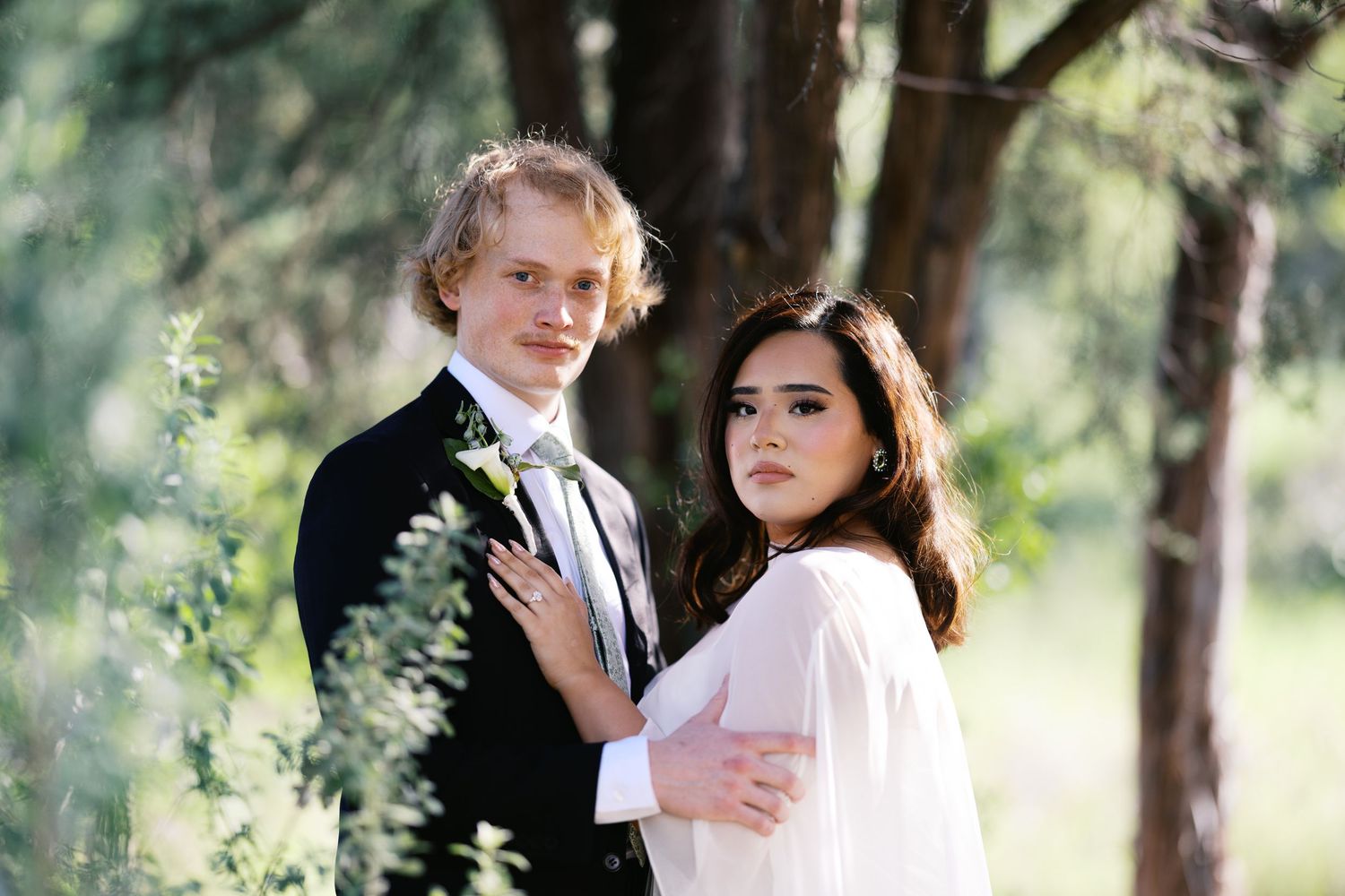 A young couple in wedding attire poses outdoors among green trees, the groom in a black suit and bride in white.