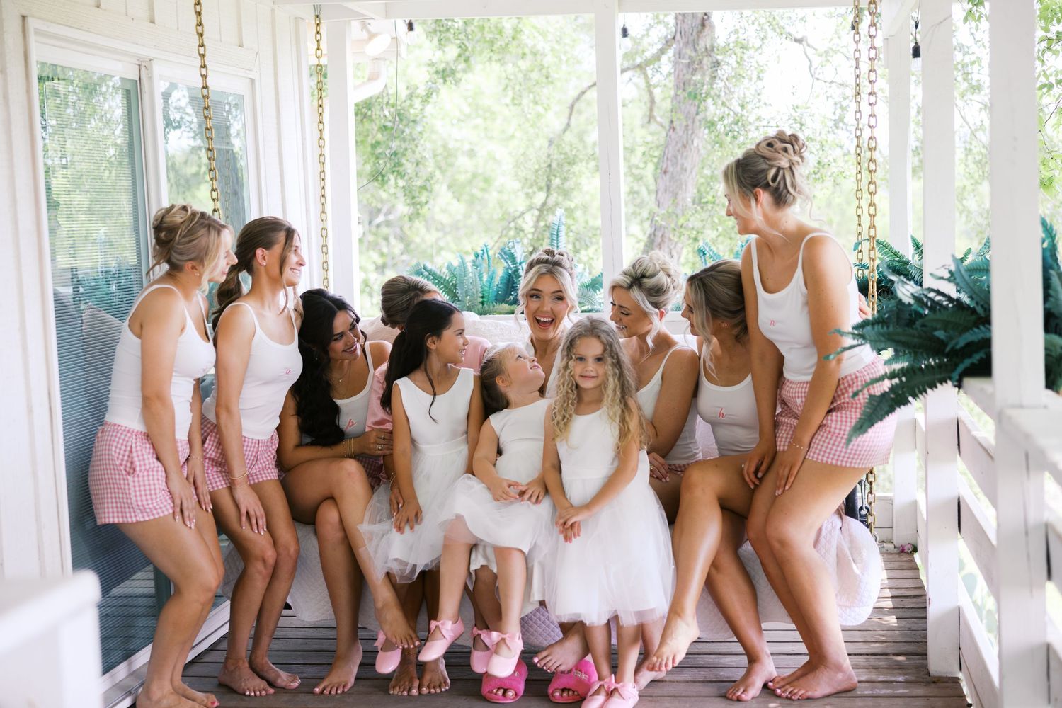 Bride surrounded by bridesmaids in white and pink outfits on a charming porch swing setting.