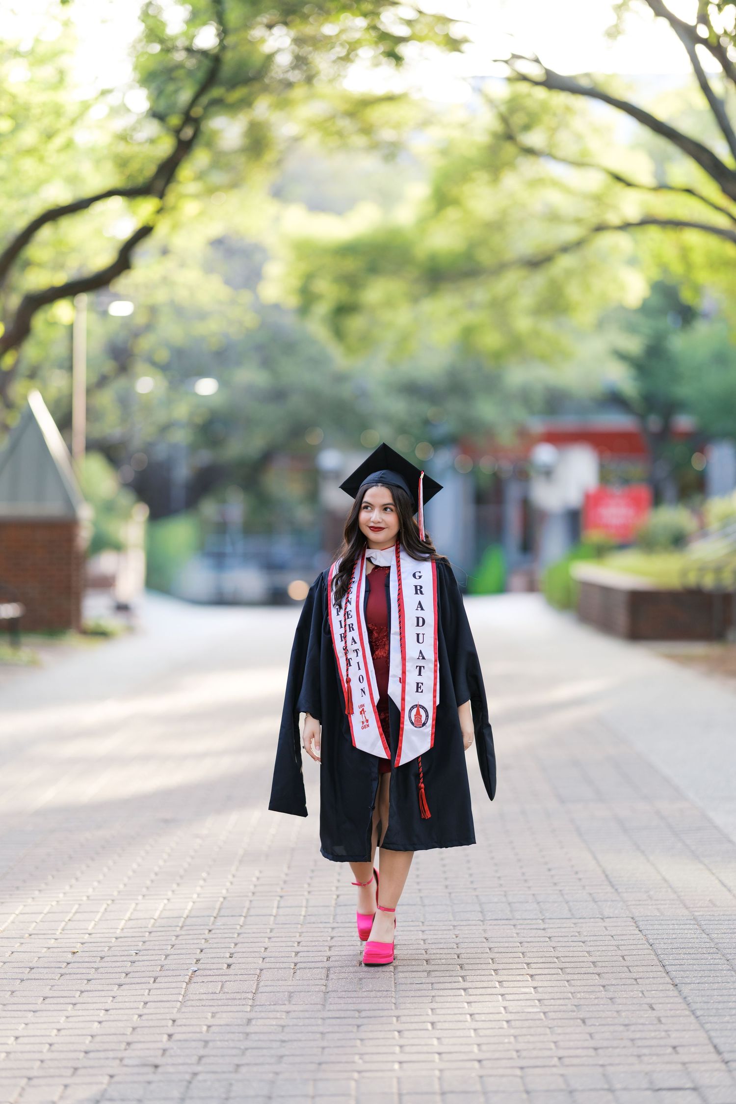 Young woman in black graduation cap and gown with red stole walks confidently on a tree-lined campus path.