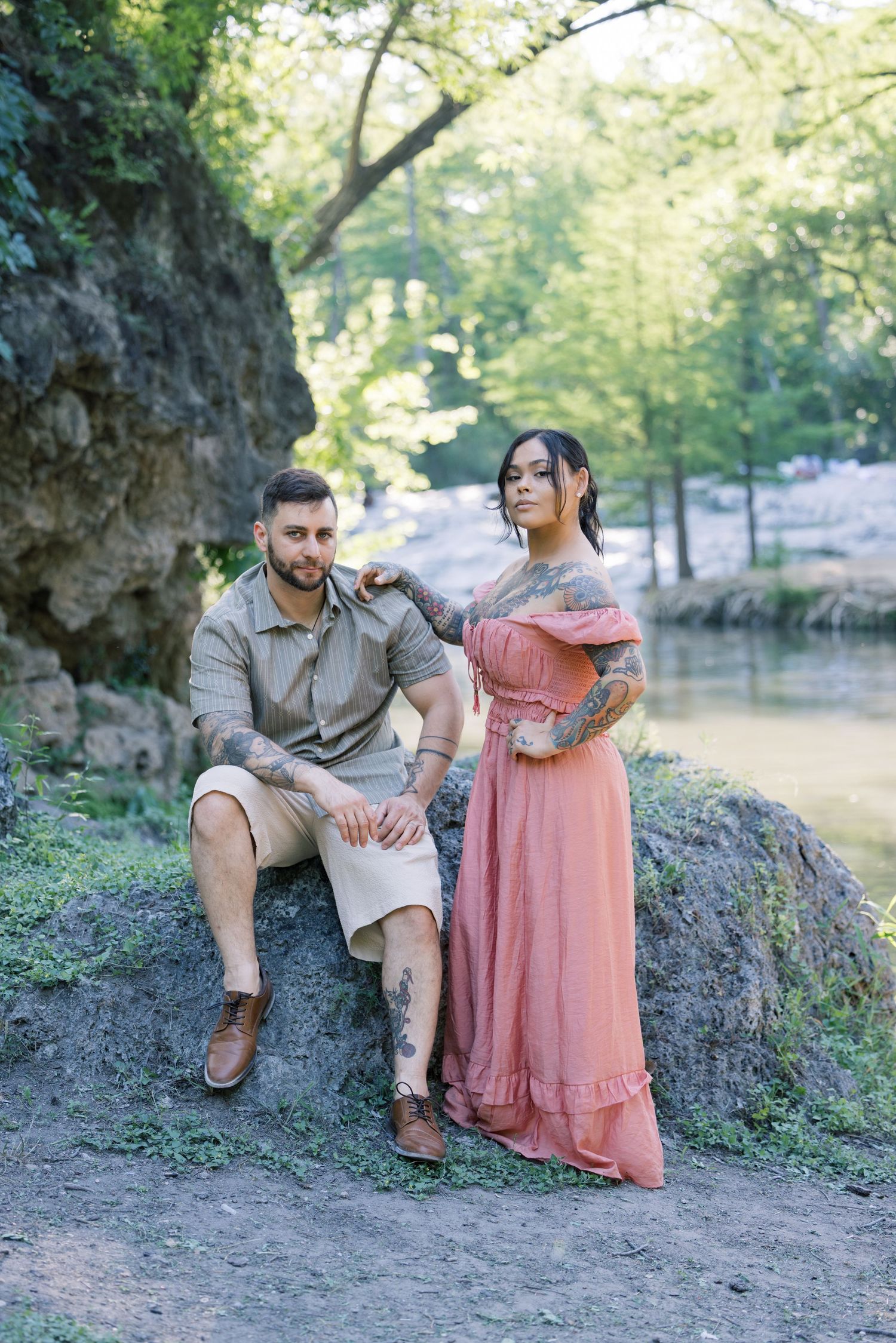 Couple posing on rocky terrain with trees and water in the background.