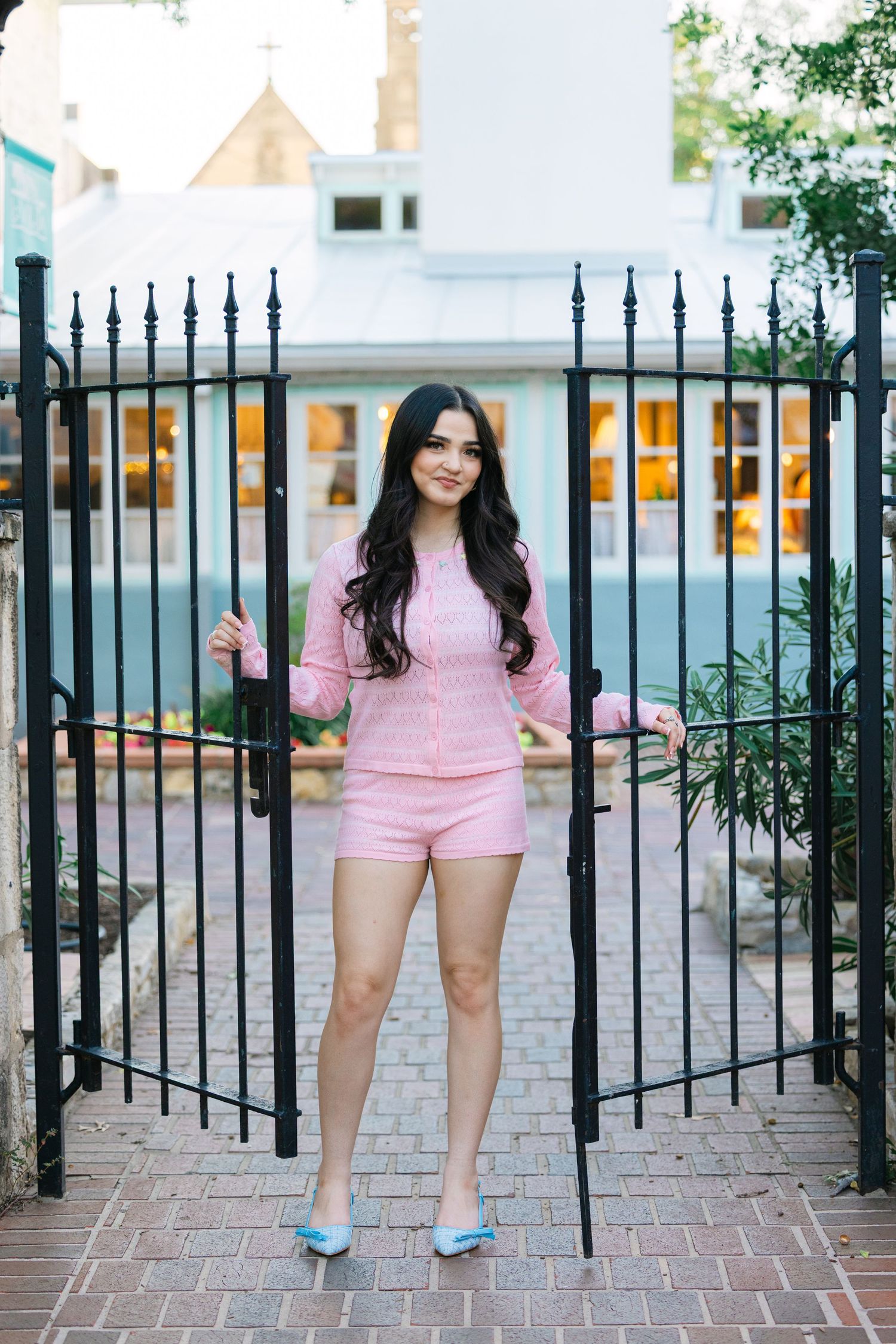A person in pink stands between ornate black iron gates on a brick pathway.