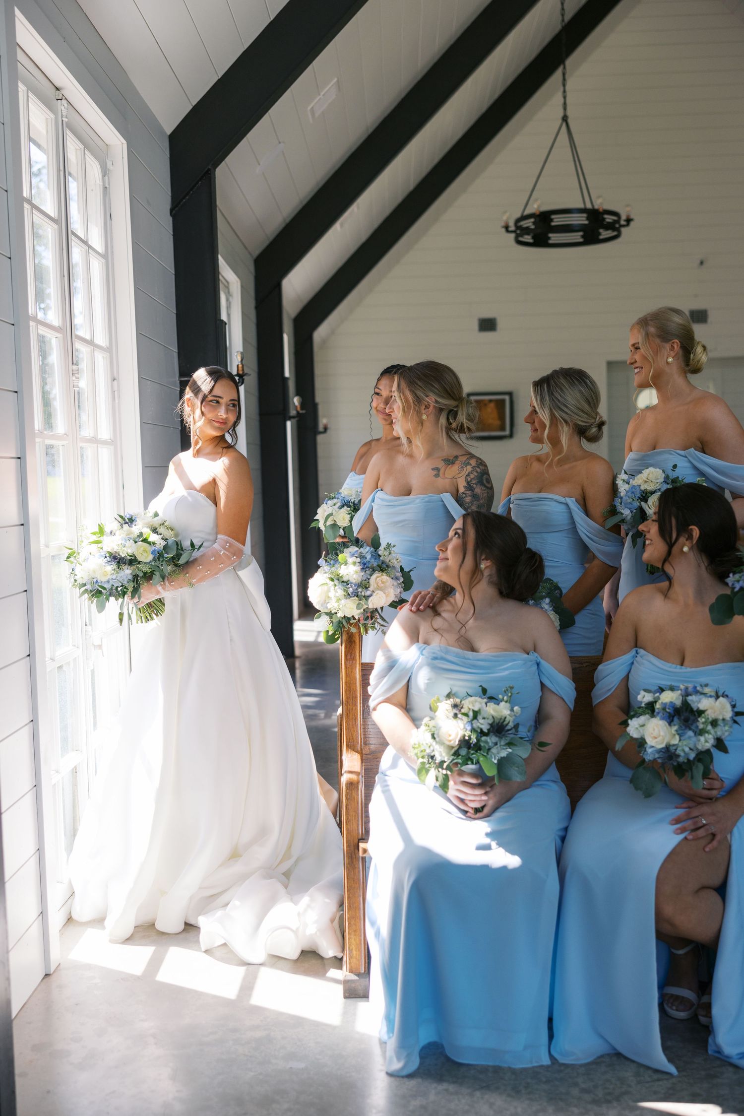 Bride in white gown poses with bridesmaids in light blue dresses near sunny window.