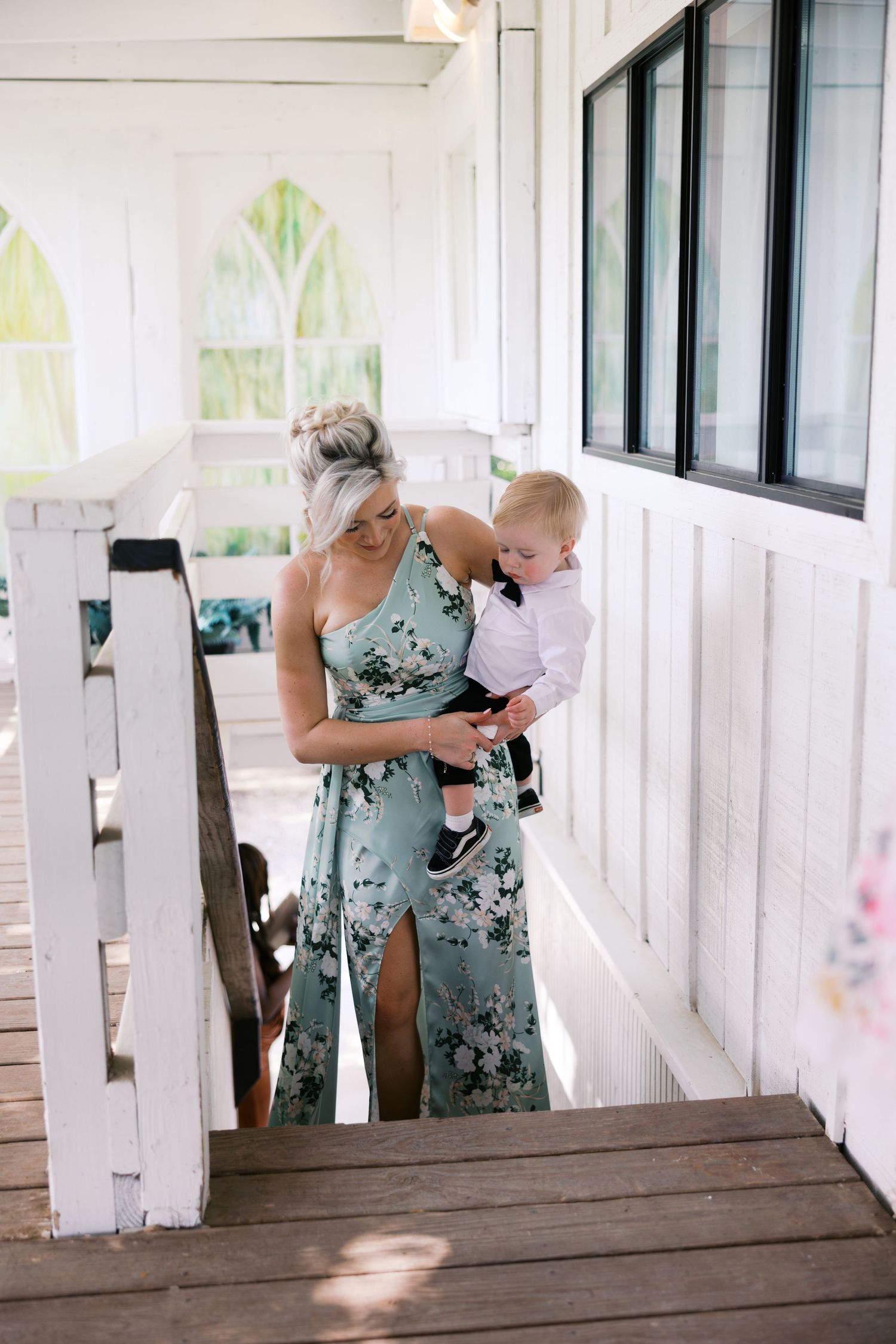 Woman in floral dress holding a baby boy on a white wooden porch at a wedding venue.