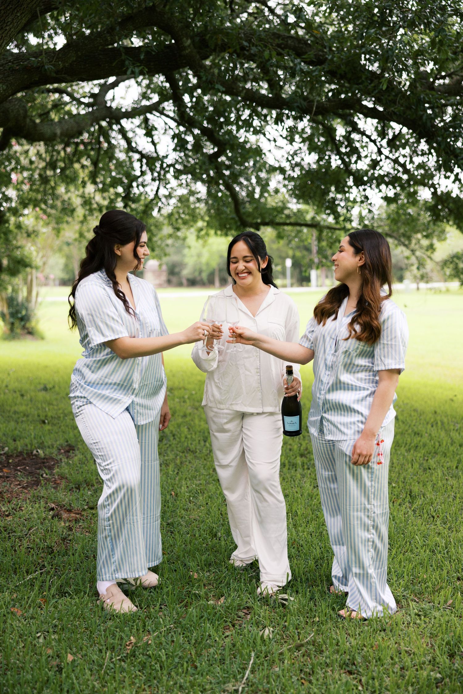 Three women in striped pajamas toasting champagne glasses outdoors under a large oak tree.