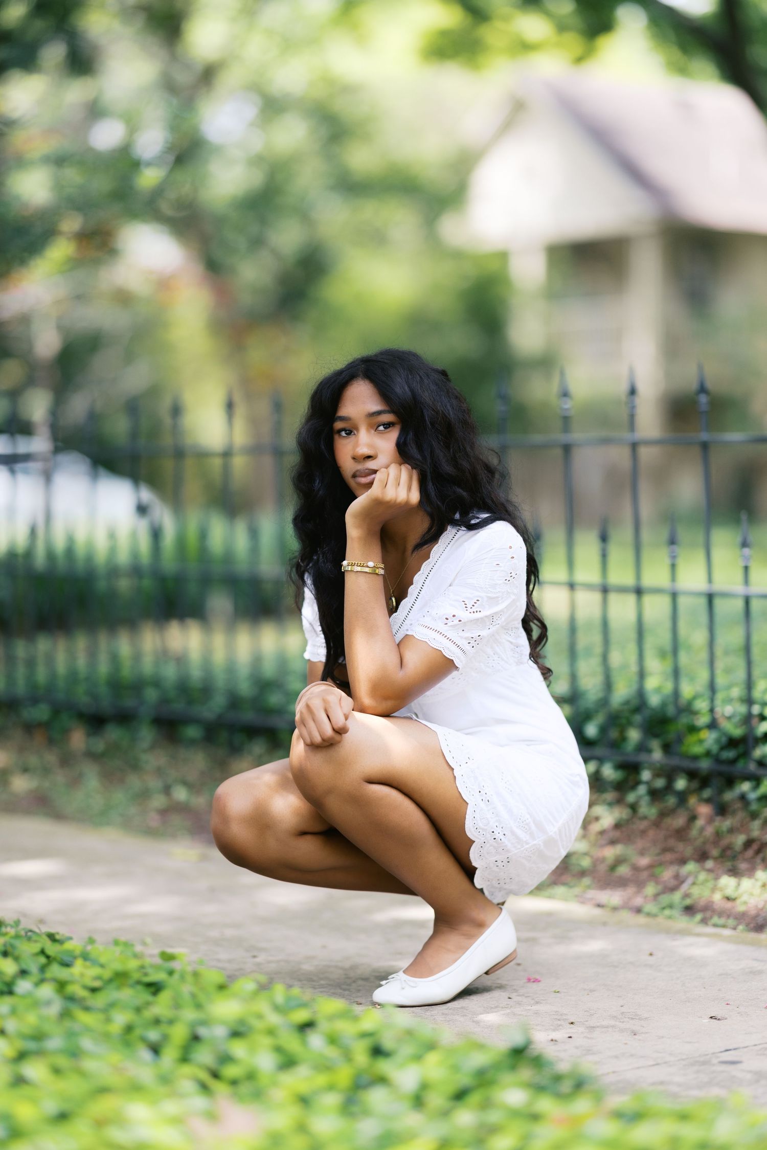 Young woman in white eyelet dress crouching gracefully on garden path beside iron fence and lush greenery.