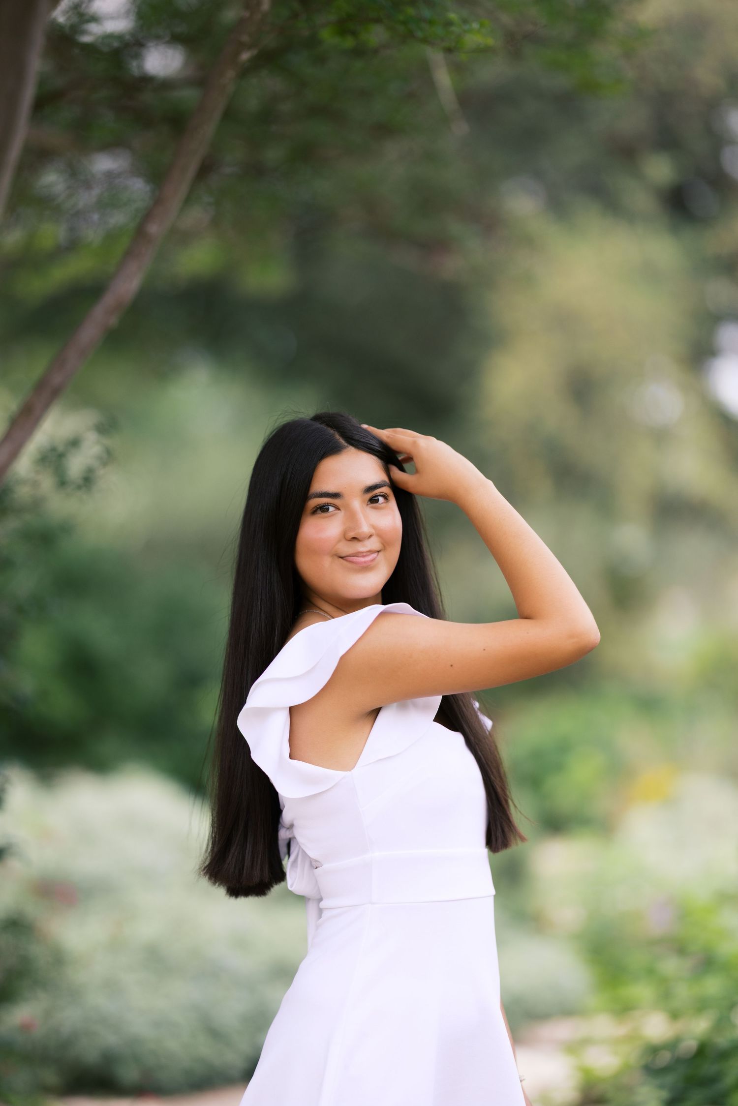 Young woman in white ruffled dress posing outdoors in a lush green garden, smiling with hand in dark hair.