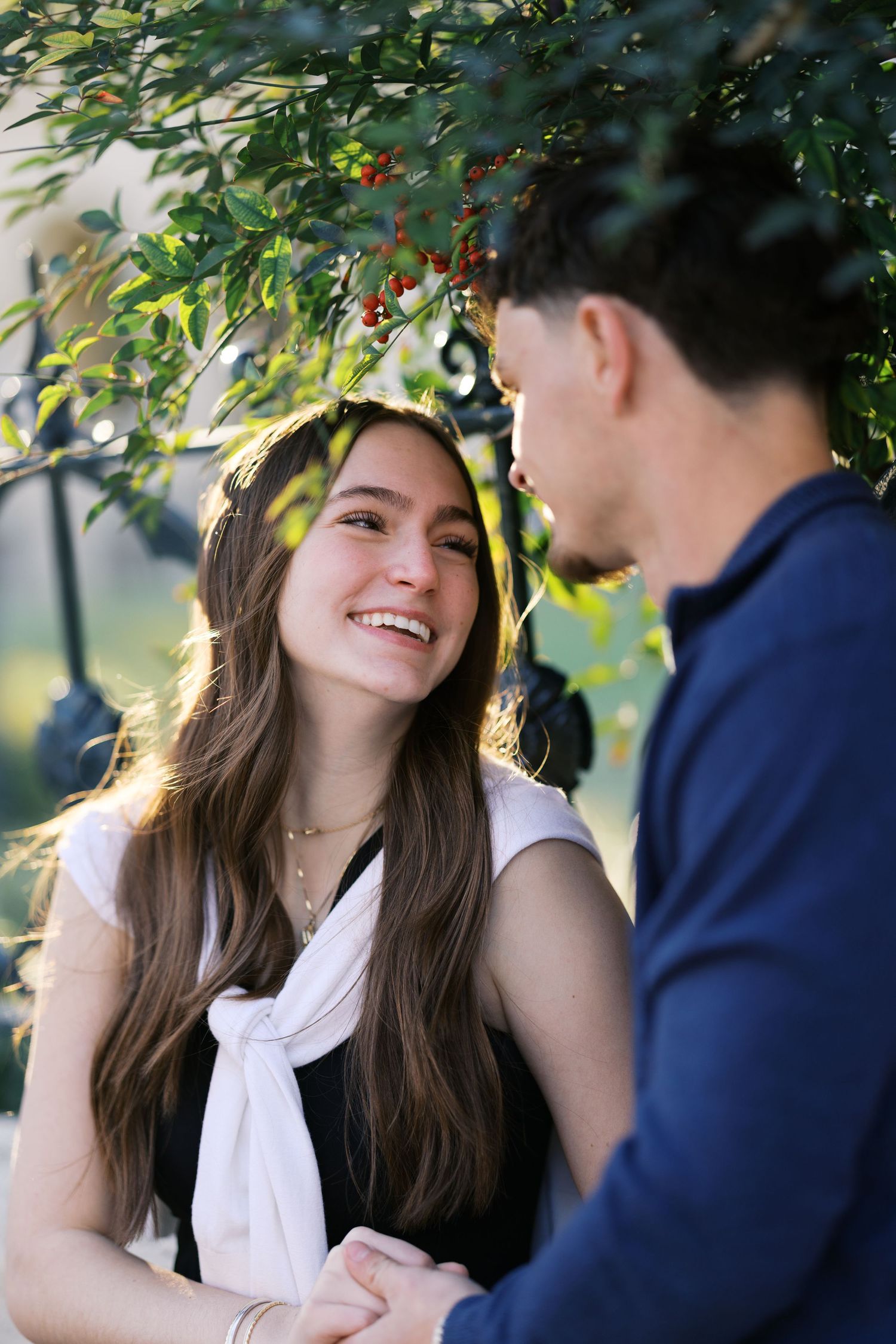 Smiling young woman with long brown hair holds hands with man beneath berry-laden branches.