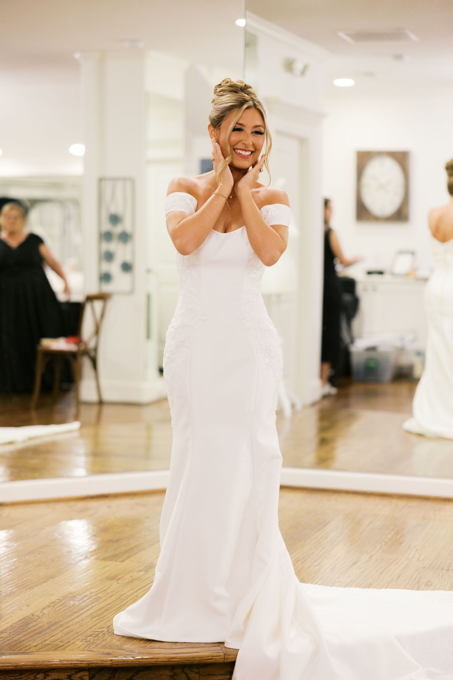 Overjoyed bride in white off-shoulder mermaid gown touching her face with excitement in bridal shop.