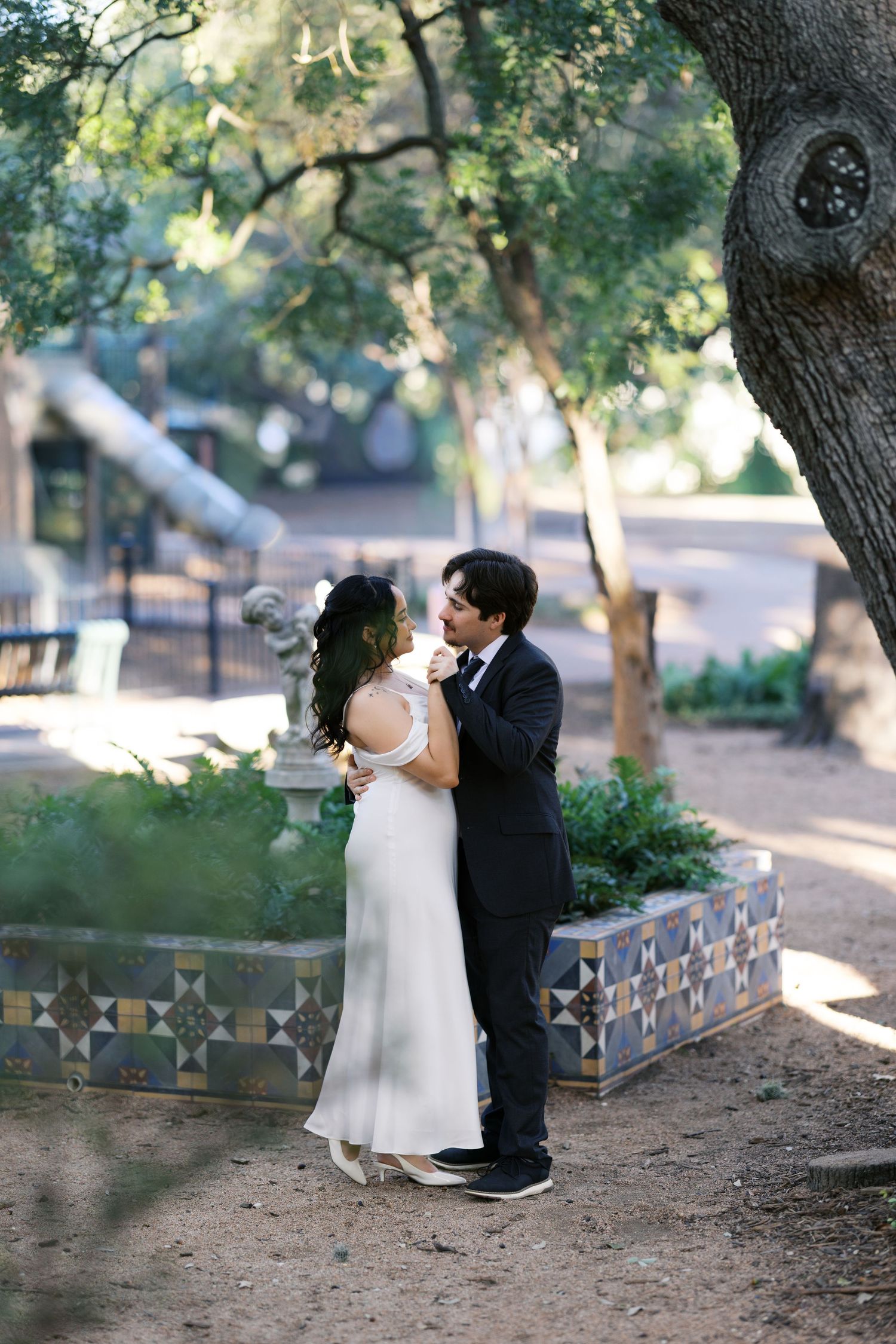 Wedding couple shares romantic moments under large trees in a garden setting.