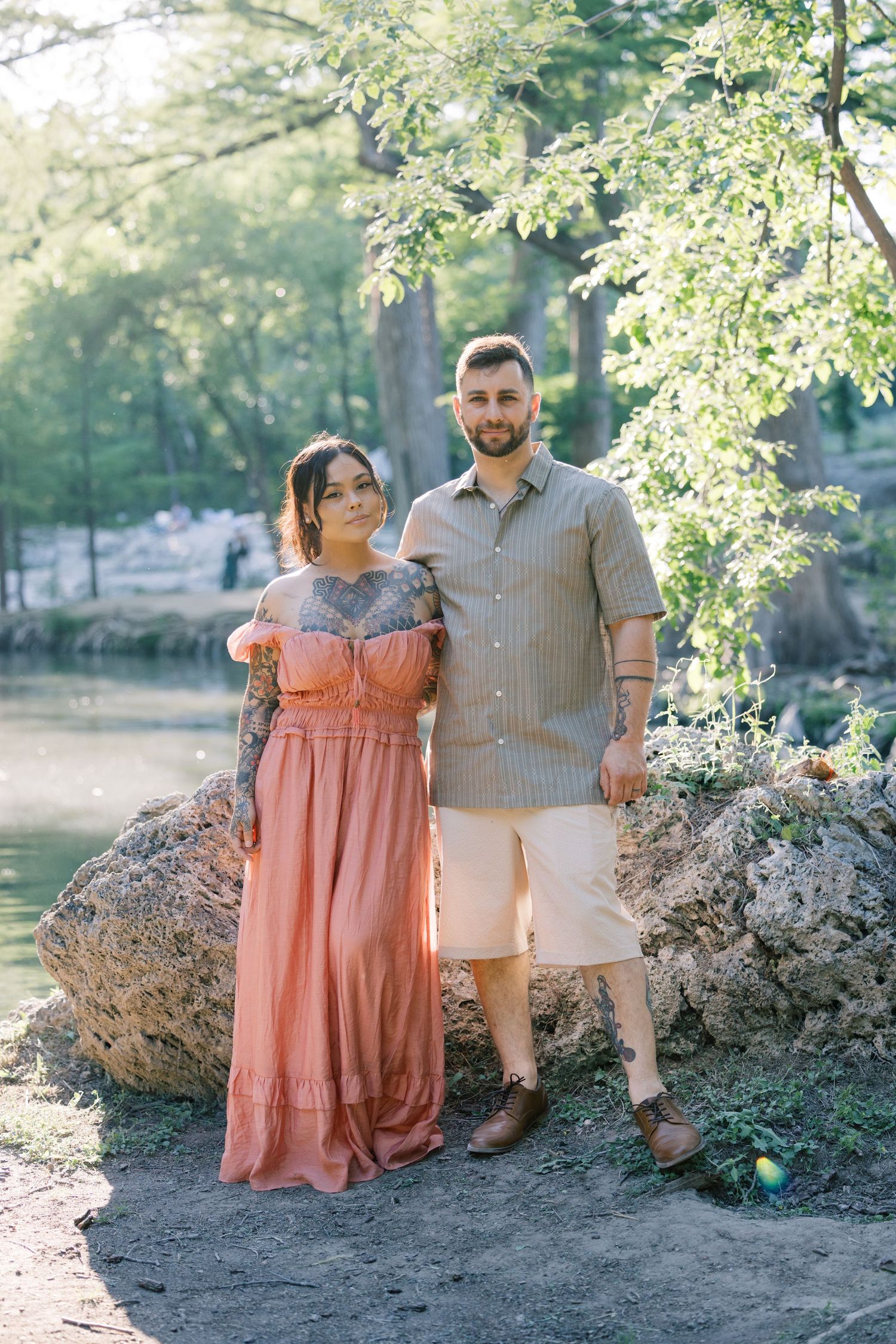 Couple in coral and beige outfits pose together against rocks and trees by a serene lake.