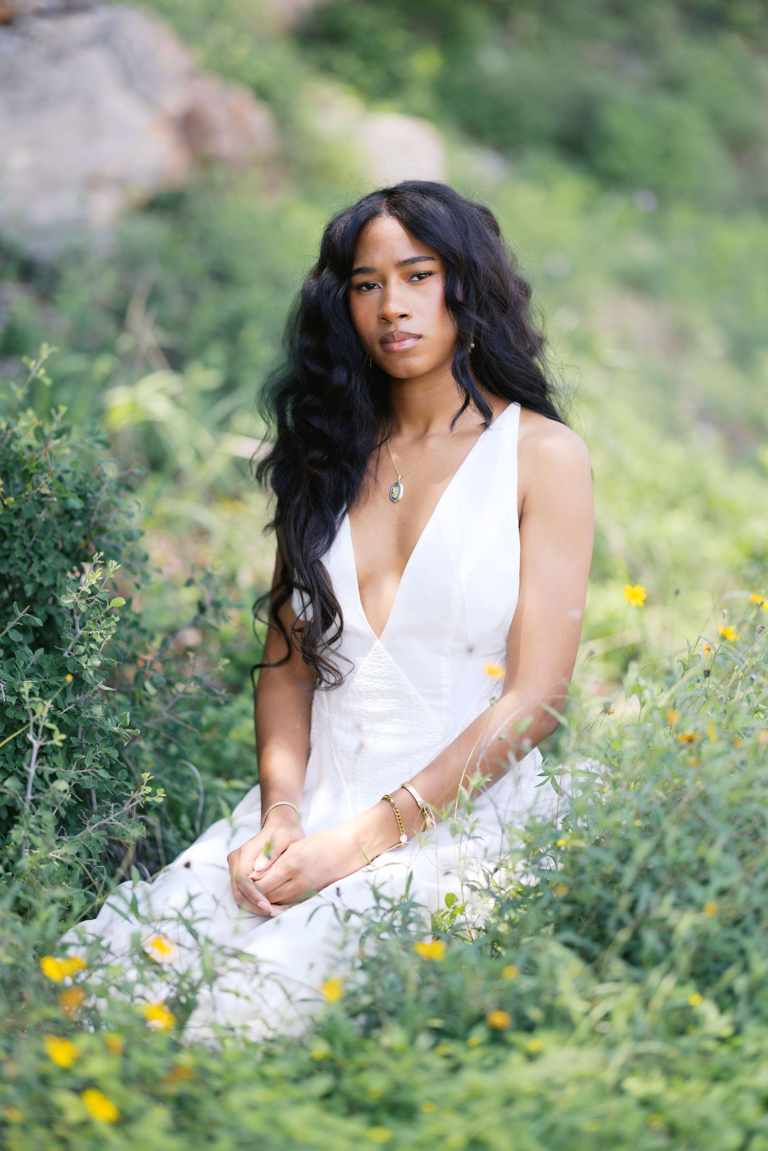 Young Black woman in white deep-V dress sitting among wildflowers on a hillside.