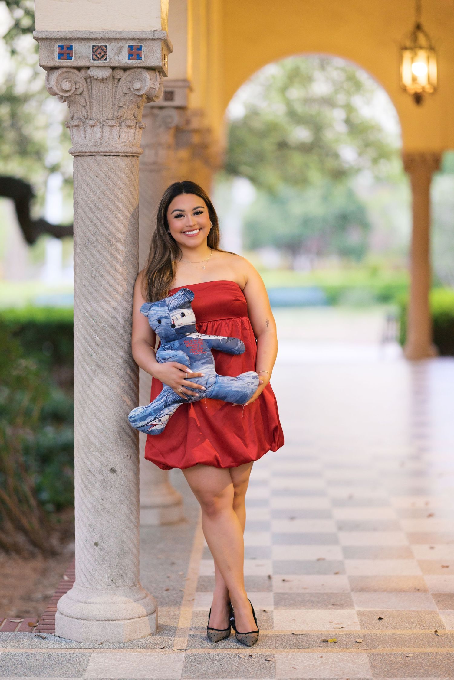 Young woman in red strapless dress holds stuffed bear while leaning against ornate column.