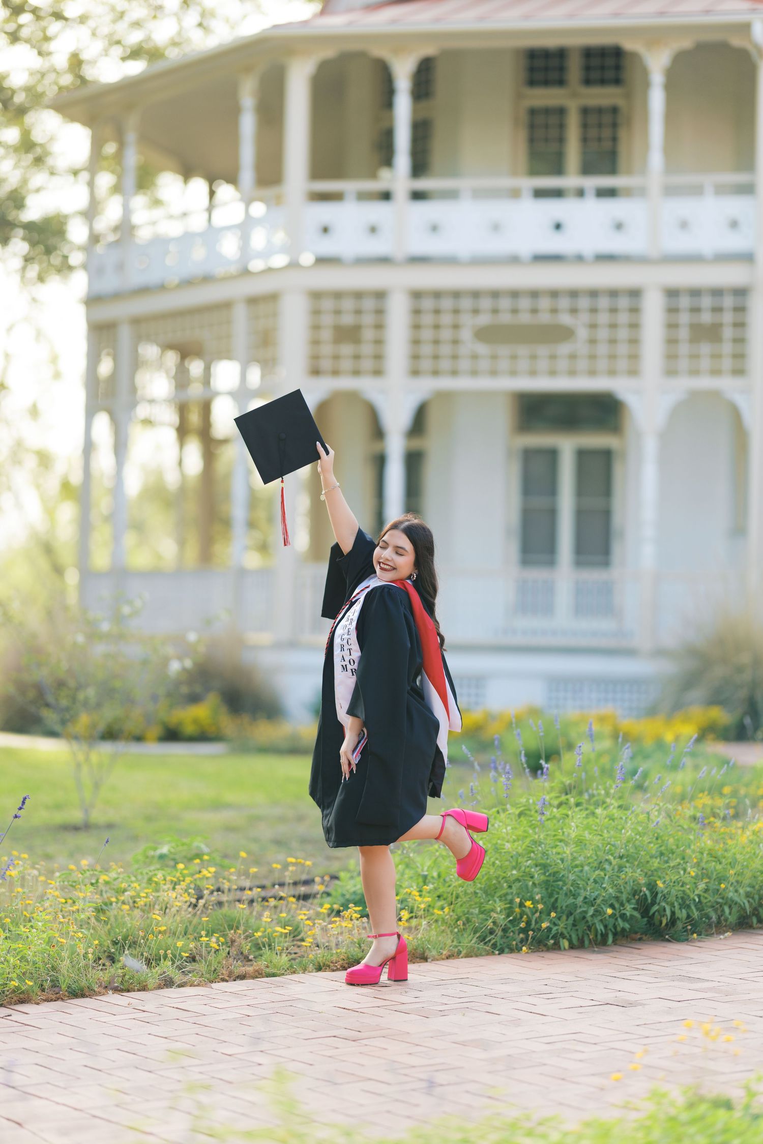 A joyful graduate in black gown and red heels raises her cap outside a white Victorian house.