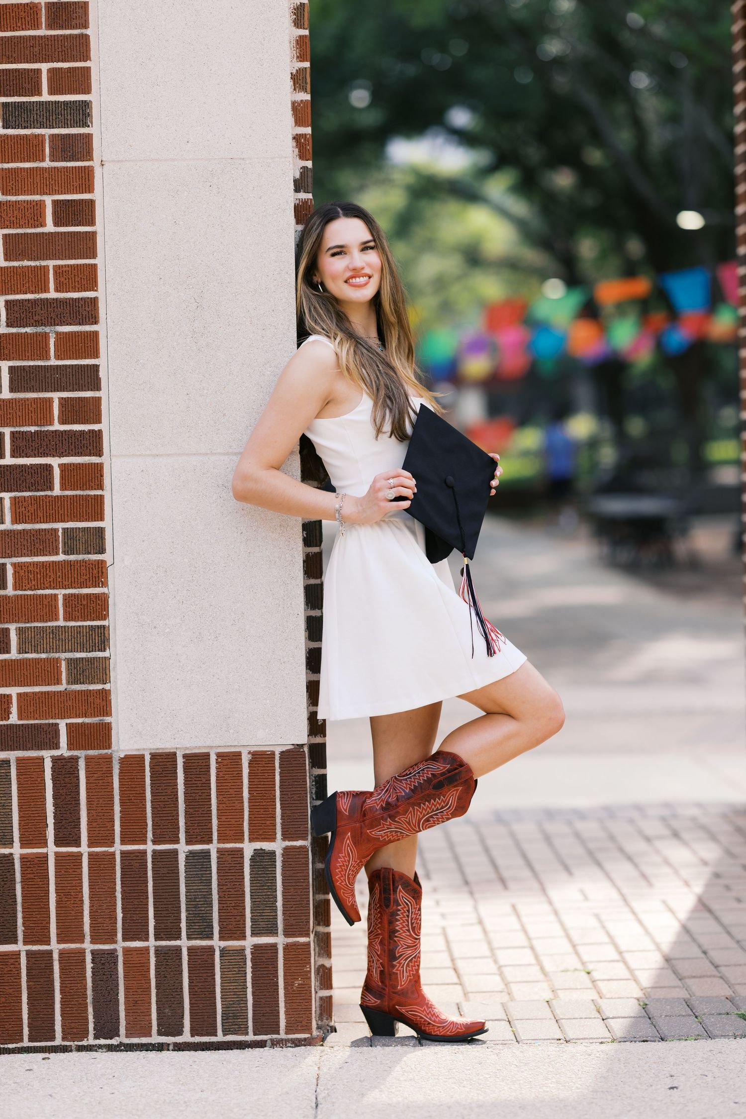 Smiling young woman in white dress and cowboy boots holding graduation cap, leaning against brick wall outdoors.