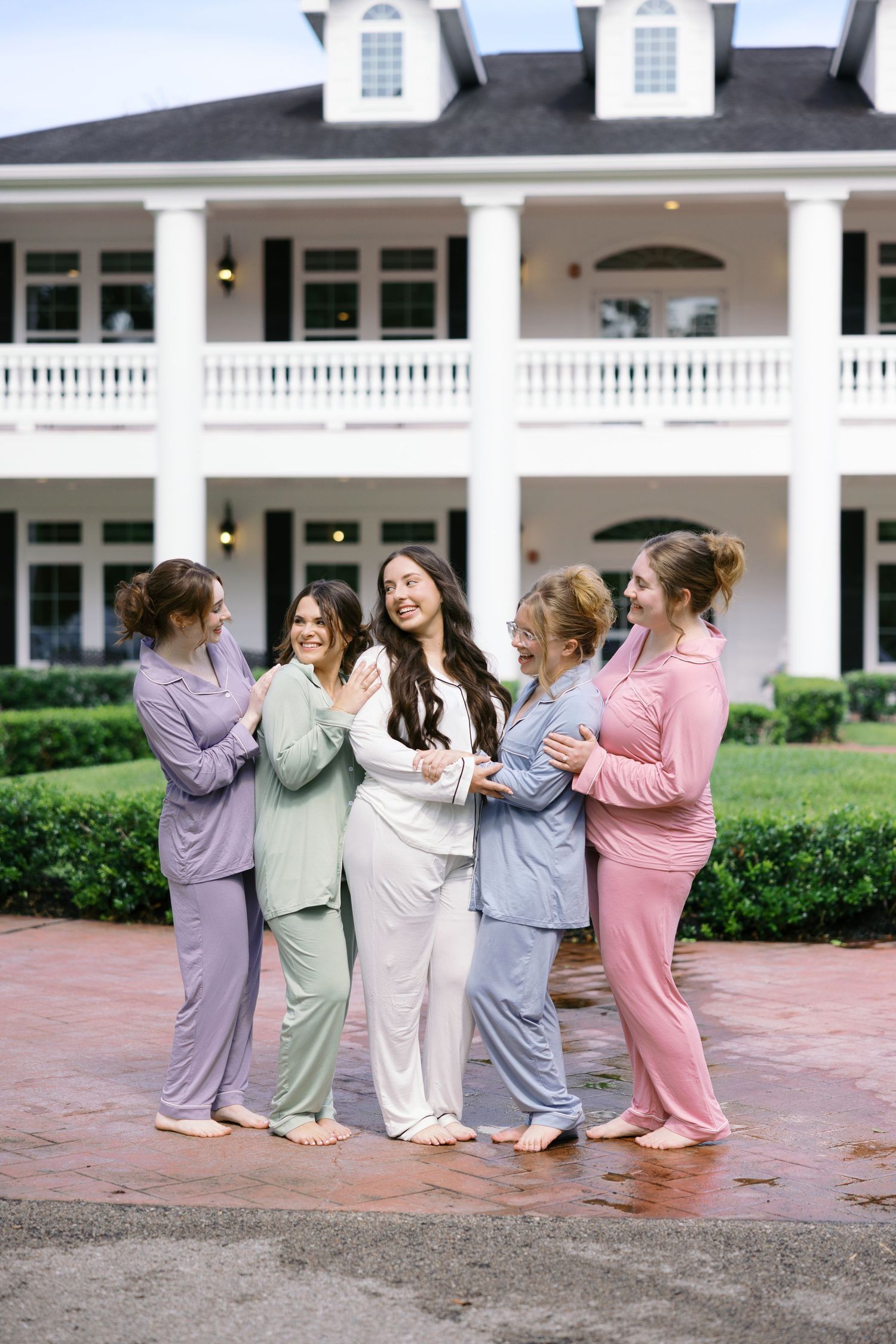 Bridesmaids in pastel pajamas doing a playful conga line pose outside a white mansion.
